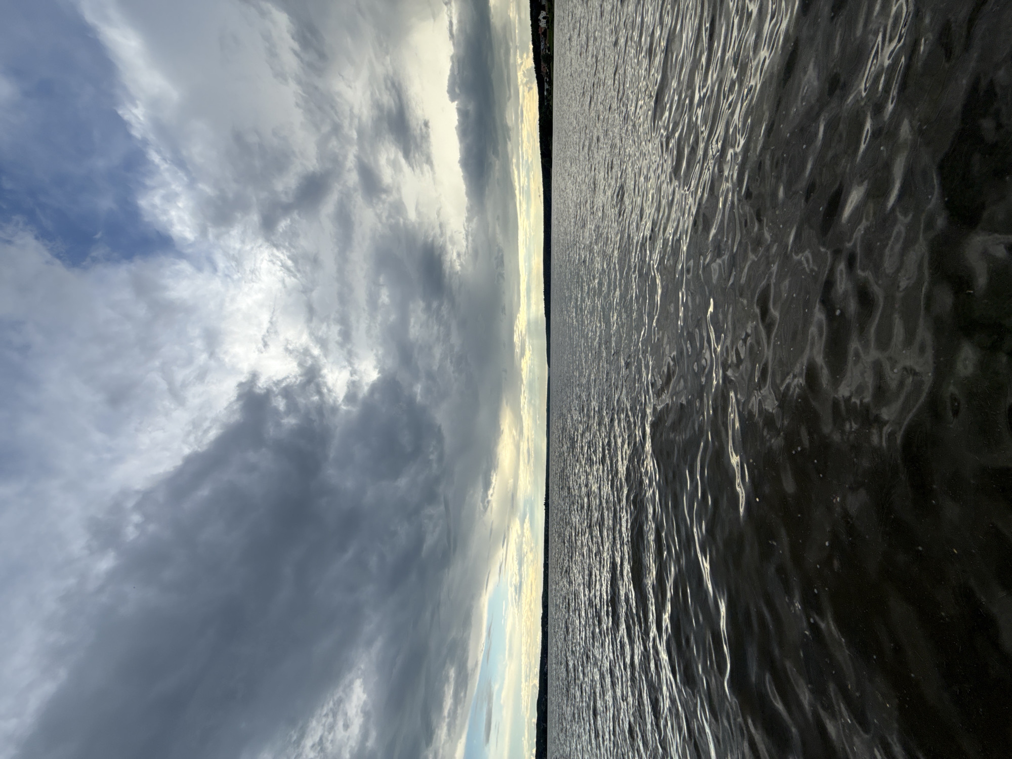 The image captures a wide expanse of water under a dramatic sky, with a distant landmass marking the horizon. The scene is presented as if viewed from on or very near the water's surface.
The upper half of the frame is dominated by a cloudy sky. Patches of bright, luminous white clouds are interspersed with darker, heavy grey cloud formations, suggesting a partly overcast or transitional weather condition. Hints of lighter blue sky are visible in some areas, particularly towards the left. The sun, though not directly visible, appears to be low on the horizon, casting bright light through gaps in the clouds and creating a luminous glow in the distance.
The lower half of the image features a vast body of water, likely a lake or reservoir in Votkinsk, Russia. The surface is covered with ripples and small waves, indicating some wind or current. Sunlight reflects strongly off the water, creating numerous bright, shimmering streaks and patterns that contrast with the darker, deeper tones of the water. The reflections highlight the texture and movement of the water.
A thin, dark strip of land with a treeline forms the horizon line, separating the water from the sky. This distant shoreline appears flat and extends across the entire width of the frame, giving a sense of vastness to the watery environment. There are no discernible people or specific objects in the frame, focusing entirely on the natural landscape. The lighting suggests it is either late afternoon or early evening, characterized by the low sun and the interplay of light and shadow on the clouds and water. The overall atmosphere is one of serene natural beauty, with a hint of an impending or recently passed weather event due to the varied cloud cover. No text is visible in the image.