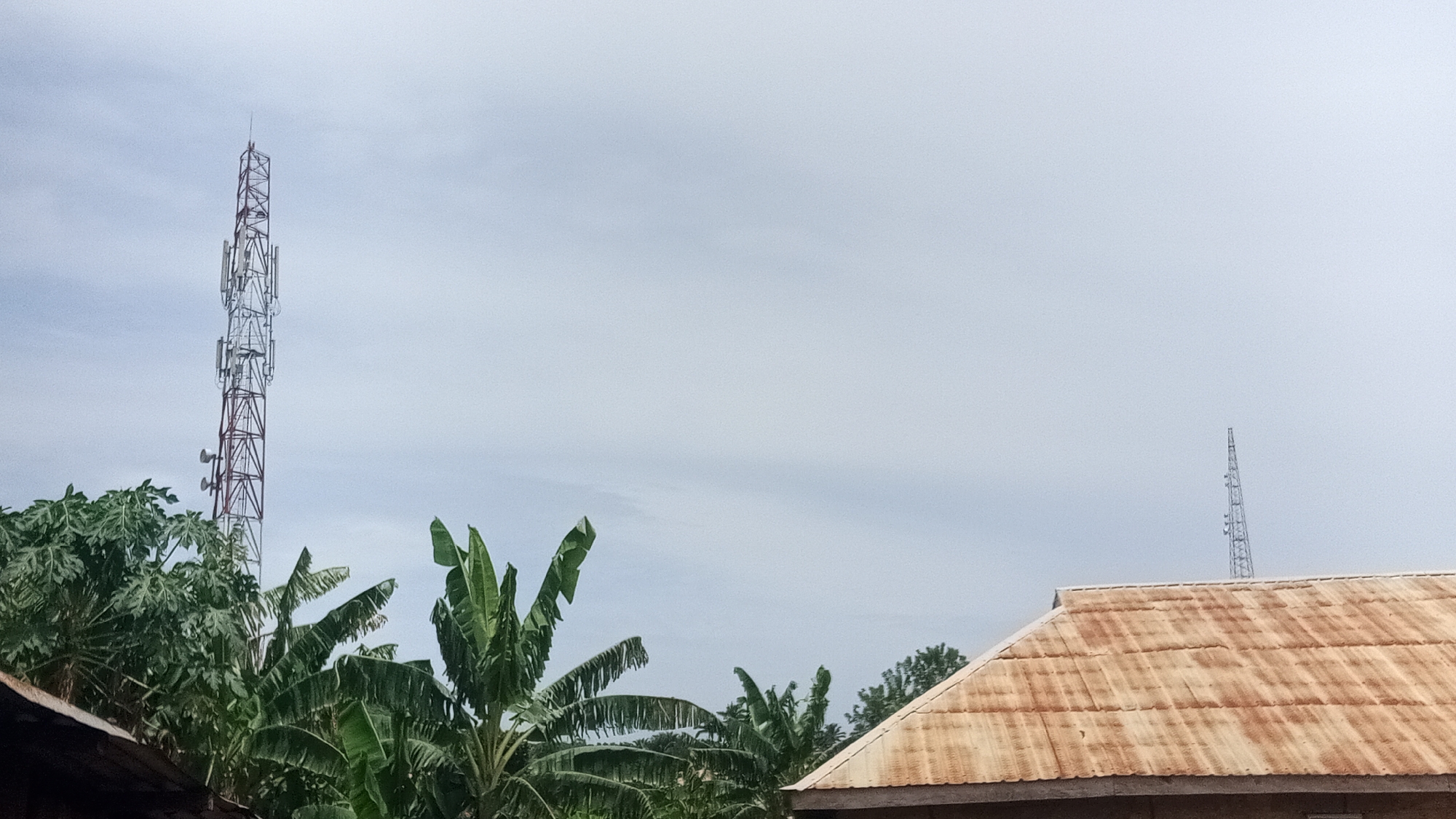 The scene depicts a daytime shot in Ikogosi, Nigeria. In the foreground, there are overgrown trees and a rusty roof with a tin-shingle-like material. Two telecommunication towers are visible, one close by and another in the far-left center of the frame. The taller one is closer to the left side of the frame and is a lattice construction painted with red and white sections, with antennas mounted on it. The other tower is more distant and appears to be of similar construction, though less distinct in detail. The sky is overcast and provides a neutral backdrop for the structures and foliage below.