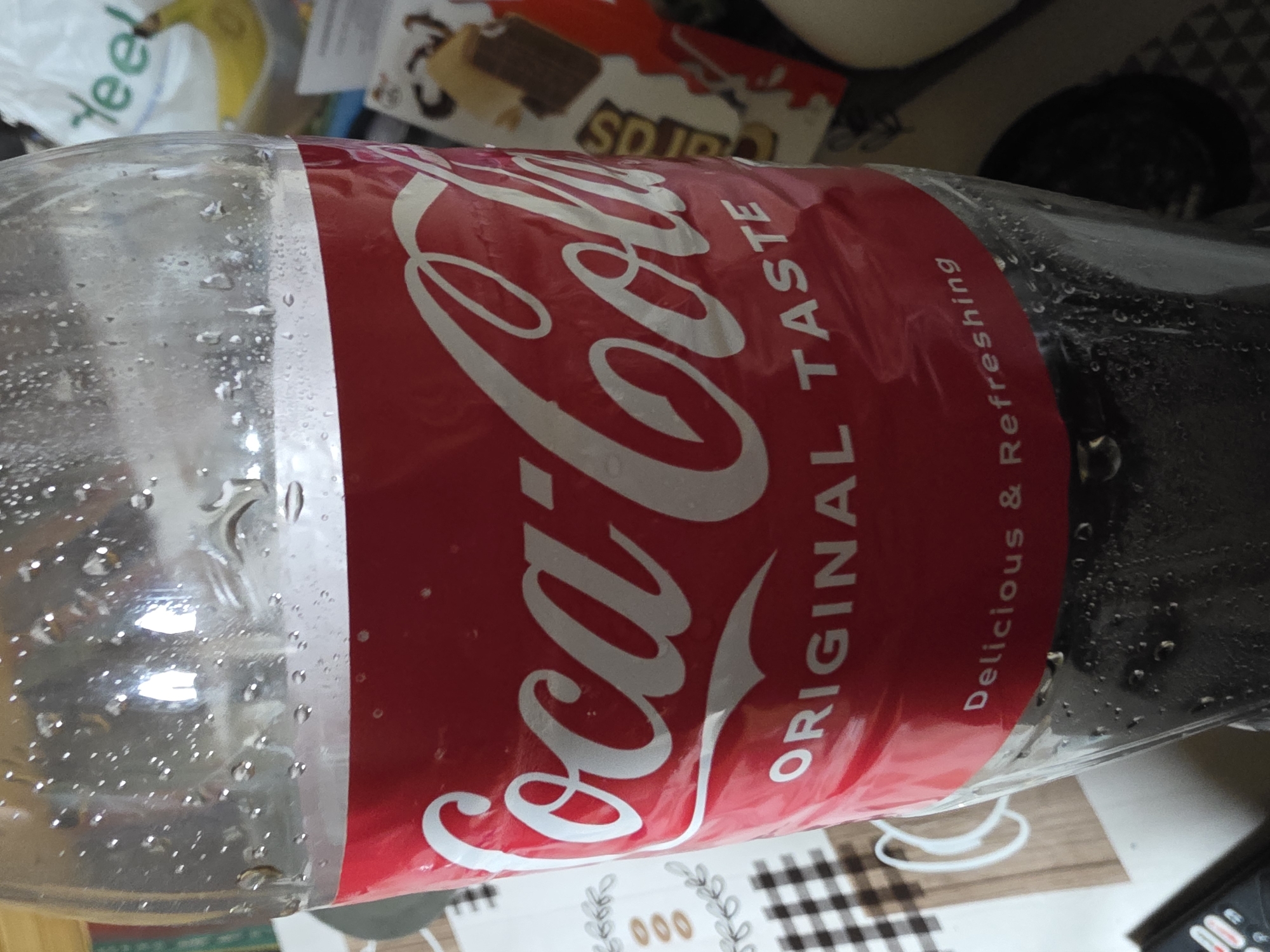 The image shows a partially empty plastic bottle of Coca-Cola lying on its side. The bottle's label is red with the Coca-Cola logo in white and the words "ORIGINAL TASTE" and "Delicious & Refreshing." Condensation droplets cover the bottle's exterior. In the background, there's a blurry view of a banana, a candy bar box labeled "SD IR," a black object, and a checkered surface. A portion of a remote control is visible in the lower right corner. There is no evidence that indicates this photo was taken in Chervena Voda, Bulgaria.