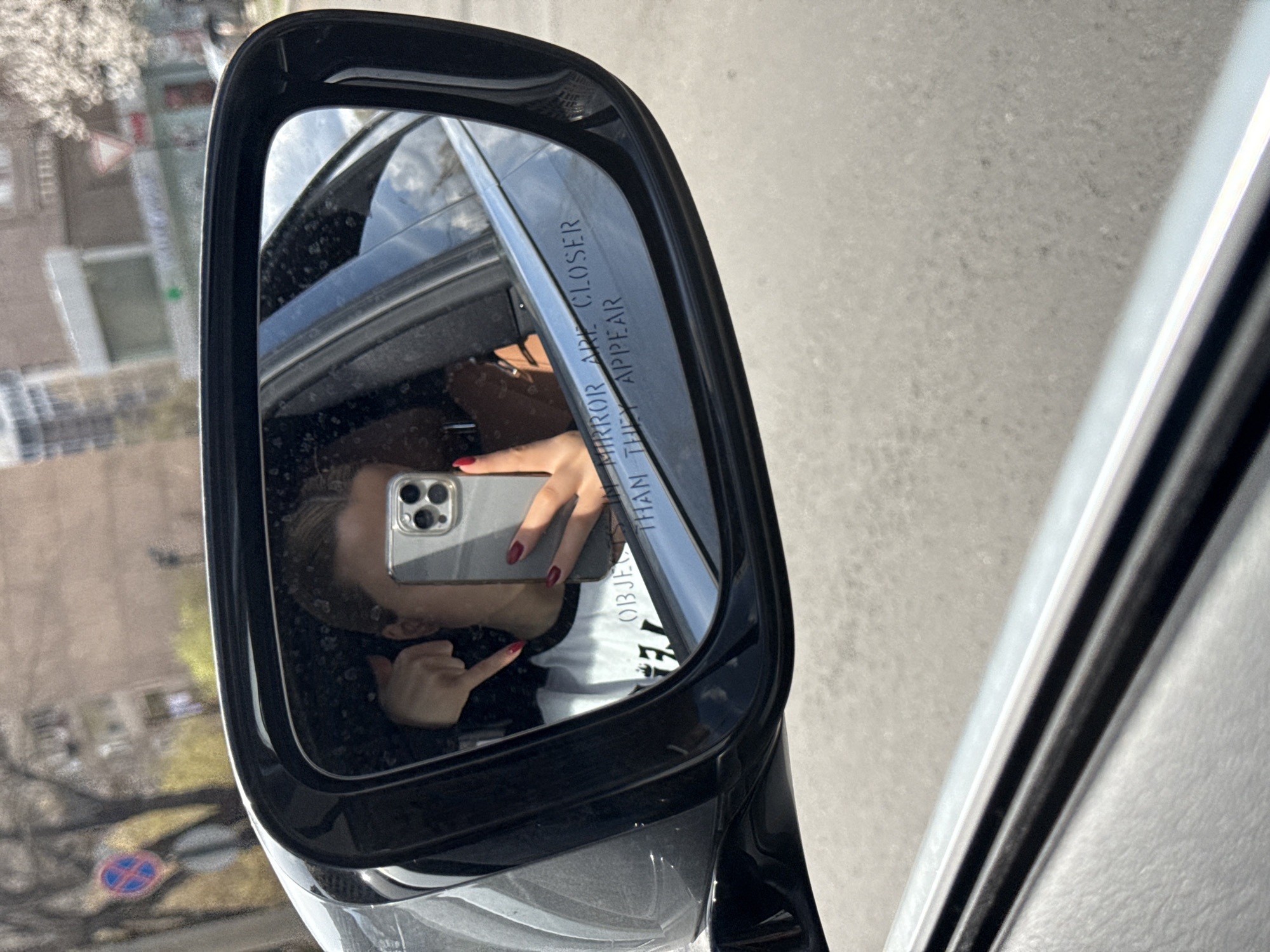 A person is taking a selfie in a car side mirror. The reflection also shows an urban environment with buildings and street signs. In the lower left corner of the mirror, a blue and red no-parking sign is visible. This suggests the location may be Yerevan, Armenia, as these colors are associated with Armenian road signs. The mirror itself has the warning "OBJECTS IN MIRROR ARE CLOSER THAN THEY APPEAR." The person is holding a gray iPhone and pointing towards themselves with their other hand. They are wearing a black jacket and a white shirt. The car appears to be parked next to a building on a sunny day.