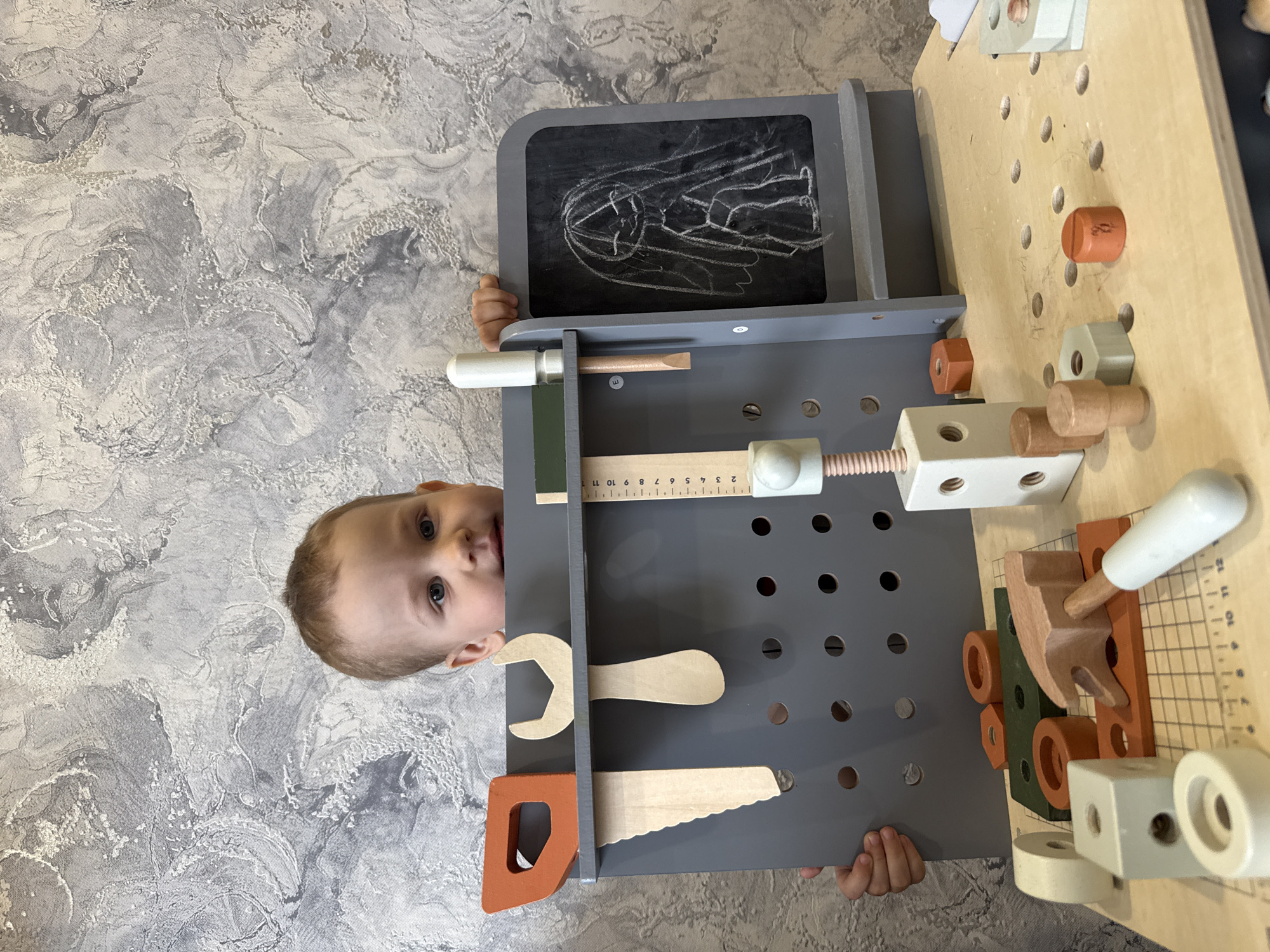 A young child is captured peeking from behind a grey and natural wood toy workbench, looking directly at the camera. The child, with fair skin, light hair, and blue eyes, has a neutral and curious expression. Only their head and portions of their hands holding the workbench are visible against a textured, grey and white patterned wall in the background.
The toy workbench is well-equipped with various play items. On the left, a small chalkboard displays a white chalk drawing of an abstract figure. Below the chalkboard, a grey pegboard section holds wooden toy tools, including a saw, a wrench, and a screwdriver. A white and grey toy vice and a wooden ruler are also attached to this section. The ruler features markings and numbers, with visible text including "8 10 11 12", "ויויויויויוייויויויו", and "2 3 4 5 6 7 8 9 10 11". The main workbench surface on the right is made of light-colored wood, perforated with many holes. Various wooden toy nuts, bolts, and building blocks in shades of orange, light green, and natural wood are scattered across or inserted into these holes, mimicking a functional workspace. The scene appears to be indoors.