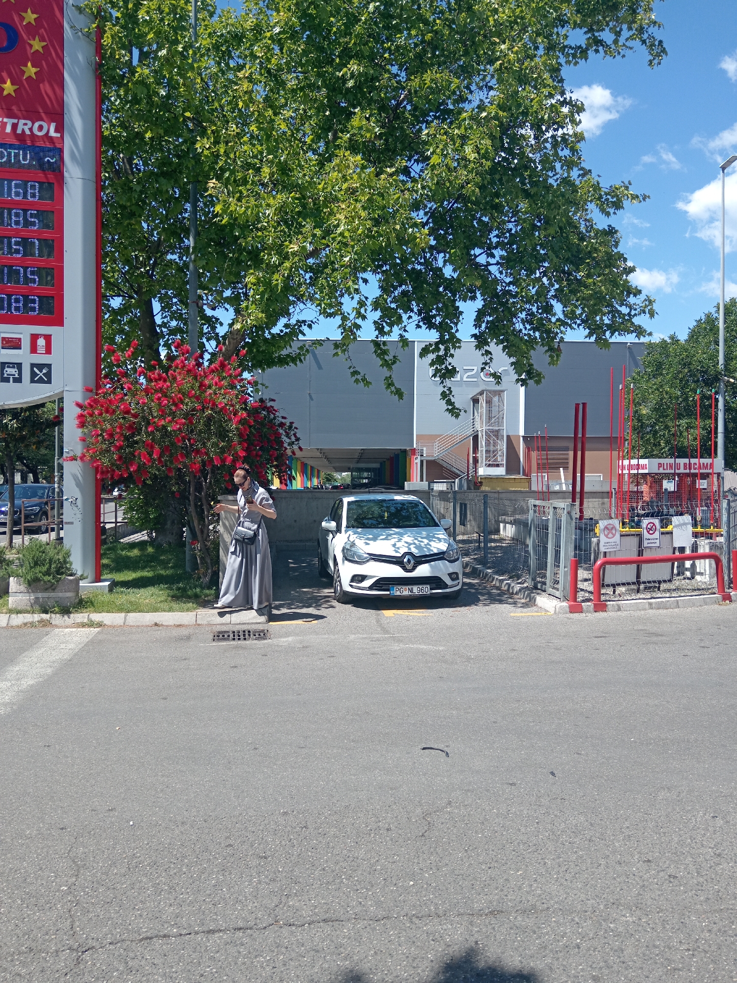 The image depicts a scene at a gas station in Podgorica, Montenegro. A woman in a long gray dress stands near a flowering tree, looking at her cell phone. Adjacent to her is a white Renault Clio with license plate PG NL 960 parked in front of the gas station. To the left, a gas price sign displays prices of 1.68, 1.85, 1.57, and 1.75. Behind the woman and the car is a large, flat-roofed building with the word "ZOR" on the upper area. Gas station equipment, including red barriers and signs related to gas cylinders (PLIN U BOCAMA), are positioned in the scene. The setting is an urban area with asphalt roads and sidewalks. The sky is blue with scattered clouds.