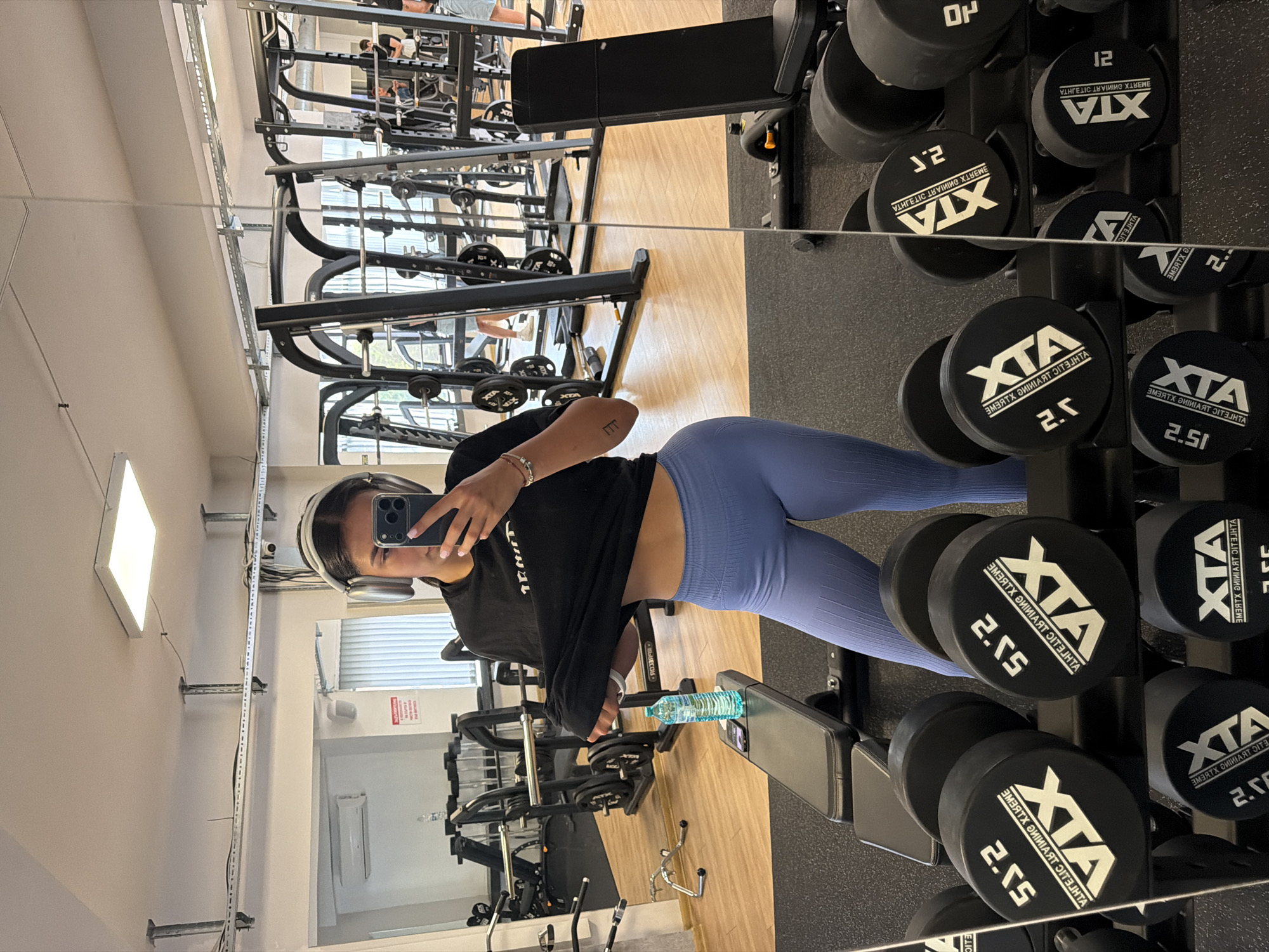 In a brightly lit gym in Gabrovo, Bulgaria, a young woman with dark hair, wearing large silver headphones, takes a mirror selfie. She is dressed in a black t-shirt, which she is lifting to reveal her midriff, and form-fitting blue leggings. She holds a smartphone in her right hand, capturing the reflection of herself and the gym equipment behind her.
The gym environment is clean and modern, with wooden flooring and a black rubberized mat in the foreground. Racks of dumbbells, each with "XTA 2.5" or "XTA 5J.2" visible on the ends, line the front of the frame. Behind the woman, a black weight bench is positioned with a bottle of water and a phone resting on it. In the background, various weight machines, including a squat rack and cable machines, are visible, along with other gym-goers, some of whom are partially obscured by equipment. The overall impression is of a functional and well-equipped fitness facility.