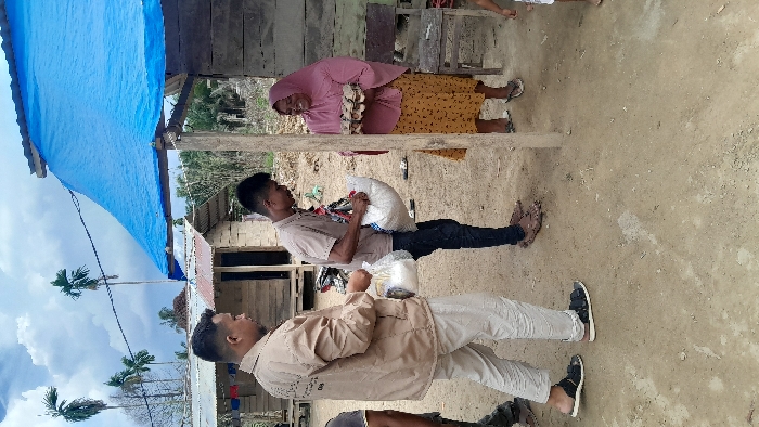 A woman in a pink headscarf and yellow skirt receives a package from a man. Another man in a tan jacket and white pants stands nearby. All three are outdoors on a dirt surface with a building behind them. A blue tarp provides overhead cover. Palm trees are visible in the background under a cloudy sky.