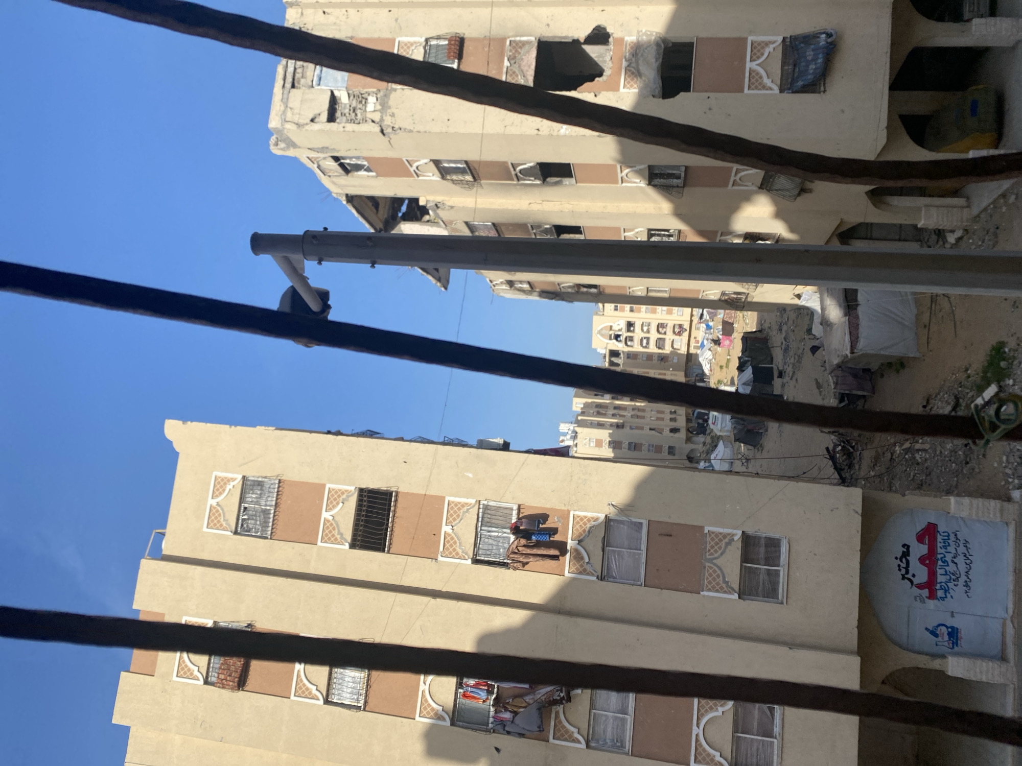The image shows a view of buildings in a city. Two main buildings are visible in the foreground, with a taller structure in the background. The foreground buildings are light brown with repetitive window designs. Some windows are open and show items inside, such as clothes or other household objects. Debris is visible on the ground between the buildings. Power lines stretch across the foreground, partially obscuring the view. A sign with Arabic writing is on the side of one of the buildings.