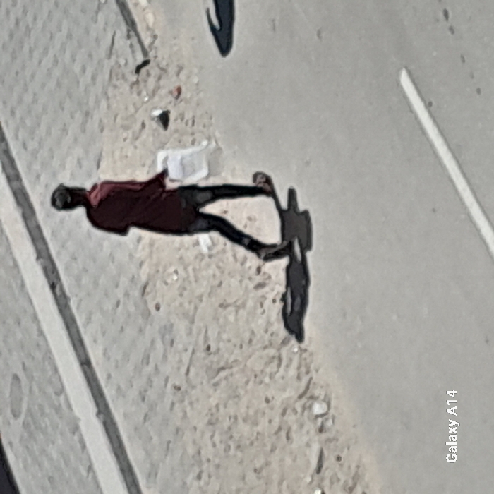 An elevated, outdoor photograph captures a lone individual walking on a street in Khan Yunis, Palestinian Territory, under bright sunlight. The person, viewed from behind, appears to be an adult wearing a dark red long-sleeved shirt, dark trousers, and flip-flops. A white object, consistent with a plastic bag, is held in the individual's right hand. The subject walks from the left to the right side of the frame, casting a distinct, elongated shadow on the ground in front and to the left. The immediate environment consists of an unpaved, light-colored ground area, possibly a dirt shoulder or undeveloped sidewalk, containing scattered debris. This area is bordered by a raised curb running horizontally across the upper portion of the image and an asphalt road surface in the foreground. A partial white painted line is visible on the asphalt road in the lower right quadrant.