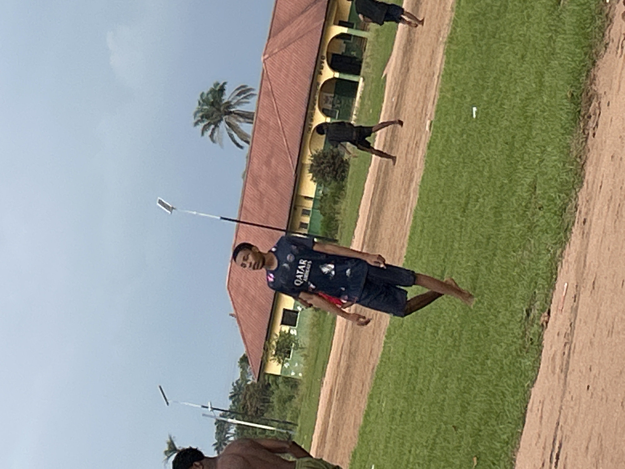The image depicts a scene in Ihiagwa, Nigeria, under a clear, possibly late afternoon sky, judging by the light. The primary subjects are three young men, likely students, engaged in outdoor activities on a grassy field adjacent to a dirt track. The man in the foreground, wearing a dark t-shirt with "QATAR AIRWAYS" branding and dark shorts, is captured mid-stride, looking towards the camera with a neutral expression. His posture suggests he might be running or posing for a photograph.
Further in the background, two other individuals are visible. One is wearing a dark shirt and shorts, also appearing to be in motion. The third person is partially obscured but seems to be wearing shorts. The setting includes a building with a reddish-brown tiled roof and yellow walls, featuring arched doorways and windows, possibly a school or administrative building. A palm tree stands to the left of the building. Two white poles, resembling streetlights or flagpoles, are present in the scene, one closer to the foreground and another further back. The overall atmosphere seems casual and active, suggesting a sports or recreational activity. The ground is a mix of dirt and well-maintained green grass.