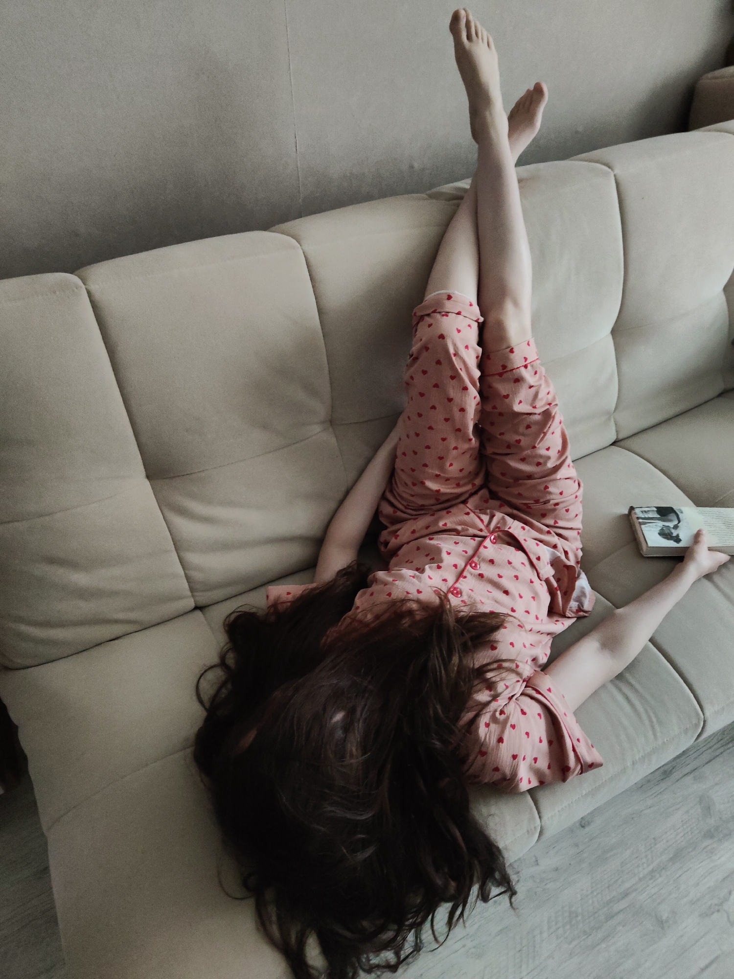 A person is depicted lying upside down on a light-colored, possibly beige or cream, sofa in an indoor setting. The individual is wearing light pink pajamas adorned with numerous small red heart patterns. Their long, dark brown, wavy hair cascades towards the floor, hanging off the edge of the sofa. Their legs are raised and crossed at the ankles, resting against the back of the sofa, with bare feet visible. In their right hand, they hold a book with a light-colored cover featuring what appears to be a black and white image or illustration and some illegible text.
The scene suggests a moment of leisure and relaxation. The sofa is a modular design with distinct cushions. The background includes a plain, light grey wall above the sofa, and the foreground shows a light wooden or laminate floor. The lighting is soft and even, characteristic of an indoor daytime setting. The person's pose conveys comfort and a casual, uninhibited state, possibly engrossed in reading or simply unwinding.
