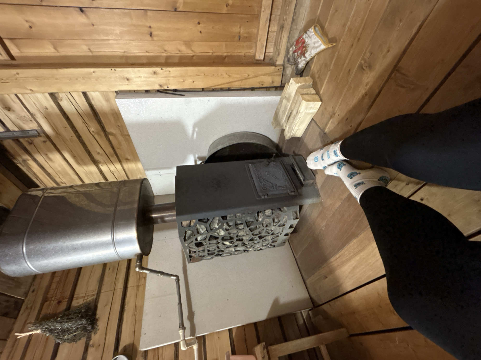 A first-person, overhead shot shows a sauna interior with wood-paneled walls and flooring. The main feature is a metal sauna stove, filled with stones. A cylindrical water tank is connected to the stove. A bundle of sauna whisks lies on the floor. A pair of legs clad in black leggings and patterned socks are present. Wood blocks and a sauna glove are on the floor. The scene appears to be inside a private sauna. The location is presumed to be Bashur, Russia.