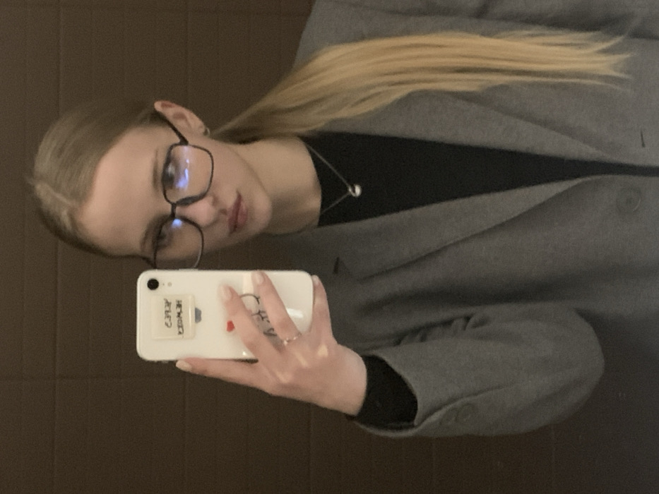 A fair-skinned woman with blonde hair in a low ponytail wears glasses and a black top under a gray coat. She is holding a white smartphone with stickers on the back, taking a selfie. Her fingernails are painted a light color. She appears to be indoors with a plain brown wall in the background. A silver necklace is visible around her neck.