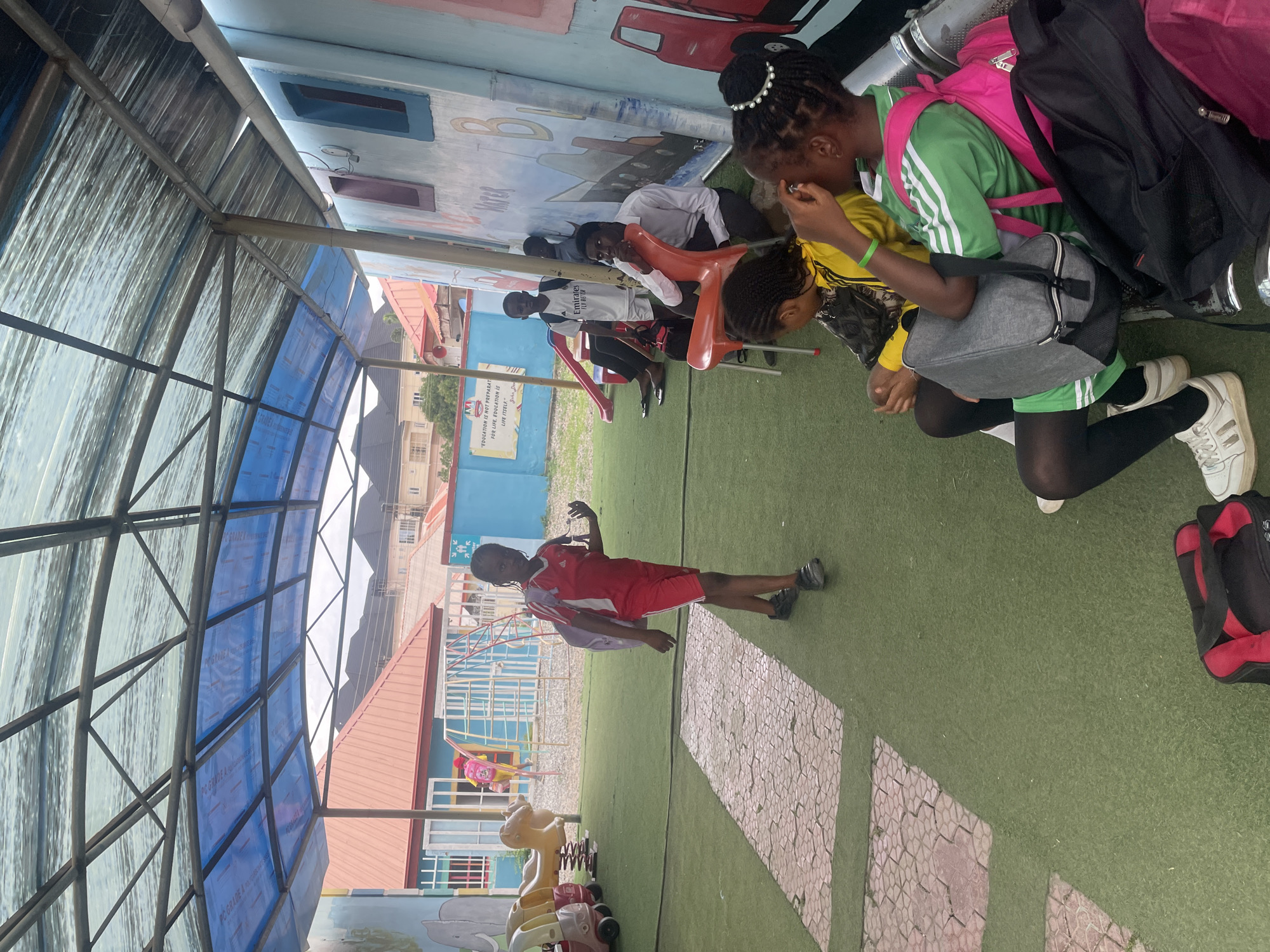 In an outdoor setting, likely a school playground in Abuja, Nigeria, several children are gathered. The scene is covered by a translucent blue and silver corrugated roof supported by metal poles. The ground is covered with artificial turf and decorated with patterned paving stones.
A group of children are seated on the floor, some wearing school uniforms. One girl in a green and white uniform with a pink backpack is prominent. Another child in a red uniform is standing, looking towards the camera. Some children appear to be listening or observing. The atmosphere seems casual and relaxed.
Visible text includes "PC GRADE A," a slogan, "Emirates FLY BETTER," and other illegible text on clothing and signs. The time of day appears to be daytime, with diffused lighting suggesting it might be overcast or under a shaded area. The overall impression is a typical school day activity in an urban Nigerian environment.