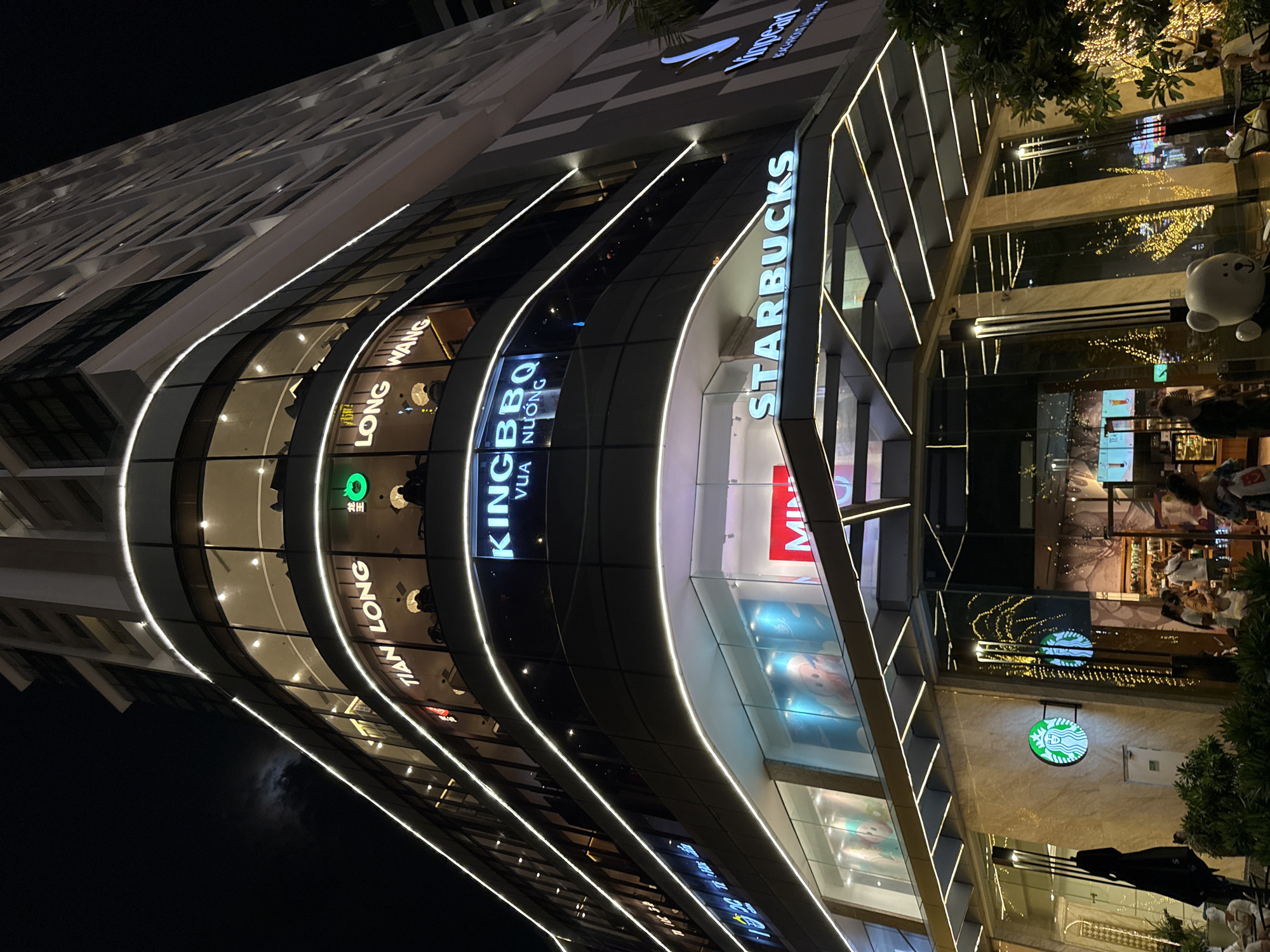 The image depicts the exterior of a modern, multi-story building at night in Nha Trang, Vietnam. The building features a curved facade with large glass windows illuminated from within, revealing various businesses. Prominent signage includes "STARBUCKS", "KINGBBQ VUA NUONG", "TIAN LONG", and "MINI". The building is adorned with white LED strip lighting that outlines the curves of the windows and architectural features. Below the illuminated upper floors, the ground level has glass storefronts. In front of the building, several people are visible, some seated at tables outside a Starbucks, while others walk by. A black umbrella stands to the left, and a white teddy bear figure is positioned near the entrance. The environment suggests a busy commercial area at night, with the bright lights of the building contrasting with the dark sky, where a faint moon is visible. The Vinpearl logo is also present on the building.