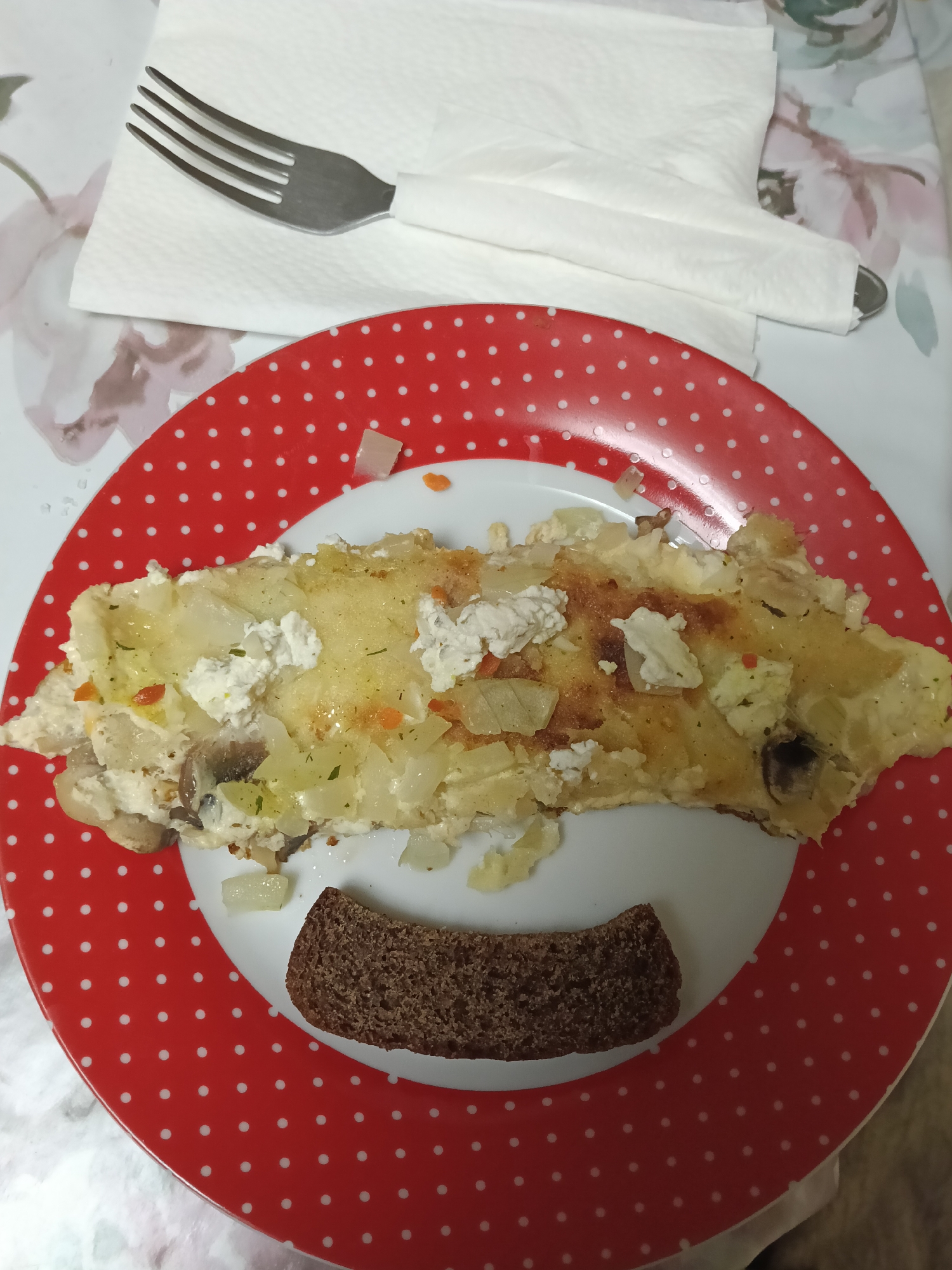 A plate of food is on a table. The plate is red with white polka dots. The food on the plate appears to be a slice of omelet with vegetables and cheese, accompanied by a piece of dark bread. A fork and a white napkin are placed to the upper left of the plate. The background appears to be a tablecloth with a floral pattern. The location is stated to be in the city of Красный Маяк, Russia.