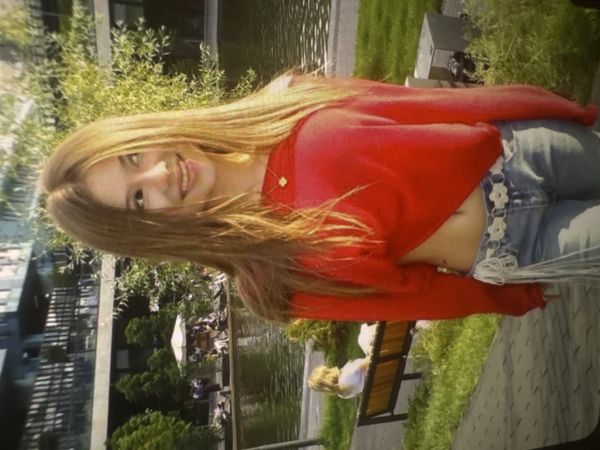 A young woman with long, straight, light brown hair smiles brightly at the camera in what appears to be an outdoor urban park setting. She is wearing a vibrant red long-sleeved crop top that exposes her midriff, light blue denim jeans, and a distinctive white belt with a floral or daisy pattern. A delicate golden necklace with a small pendant, possibly a butterfly or heart, is visible around her neck. A small, dark tattoo can be seen on her left side, just above the waistband of her jeans.
The background features a body of water, possibly a river or pond, reflecting the sky and surrounding greenery. Lush green bushes and trees frame the scene, suggesting a warm and sunny day. In the distance, modern buildings with glass facades are visible, indicating an urban environment. Another individual, with light hair, is seen sitting on a bench near the water's edge, facing away from the camera. The overall impression is one of a cheerful moment captured in a pleasant, well-maintained public space. There is no visible text in the image.