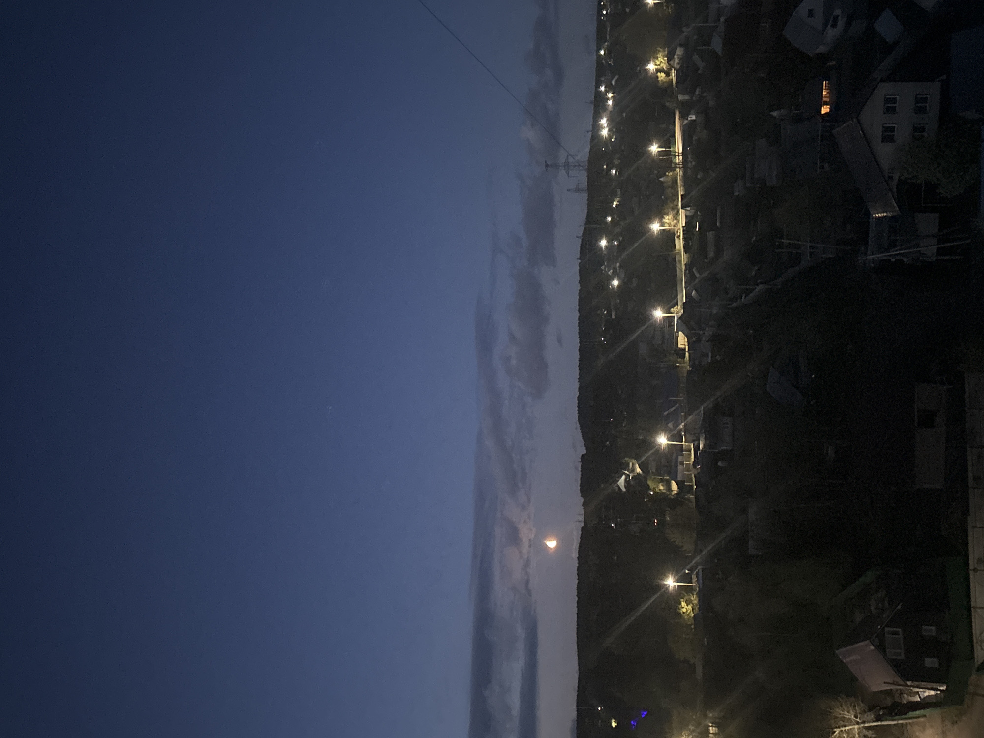 The image depicts a night scene in Chaikovsky, Russia. On the left, a dark blue sky transitions to a vertical band of clouds. To the right of the clouds, a hillside is visible, dotted with residential buildings and illuminated by streetlights. A powerline is suspended above. A crescent moon is visible within the cloud band. Buildings with illuminated windows can be seen among the dark houses.