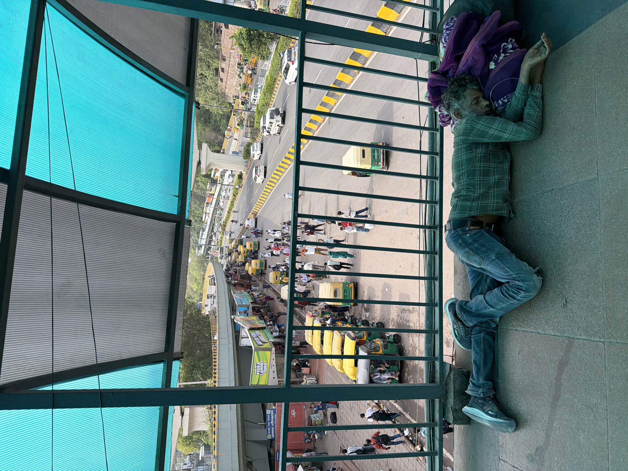 The image depicts a street scene in Delhi, India, viewed from an elevated pedestrian walkway. In the foreground, a man is sleeping on the ground, covered by blankets and a bag. He is lying on his side, wearing a plaid shirt and blue jeans, with his head resting on a purple blanket. His feet are clad in sneakers.
Below the walkway, a busy street is visible with multiple lanes of traffic. Various vehicles, including cars, auto-rickshaws (also known as tuk-tuks), and buses, are present. The auto-rickshaws are predominantly yellow and green. Pedestrians are seen walking along the sidewalk and near the vehicles. Structures indicating a bus station or market area are visible along the street, with shops and people gathered. An elevated road or flyover is also a prominent feature of the urban landscape. The walkway itself is covered by a canopy made of translucent blue and grey panels. The scene appears to be during daylight hours.