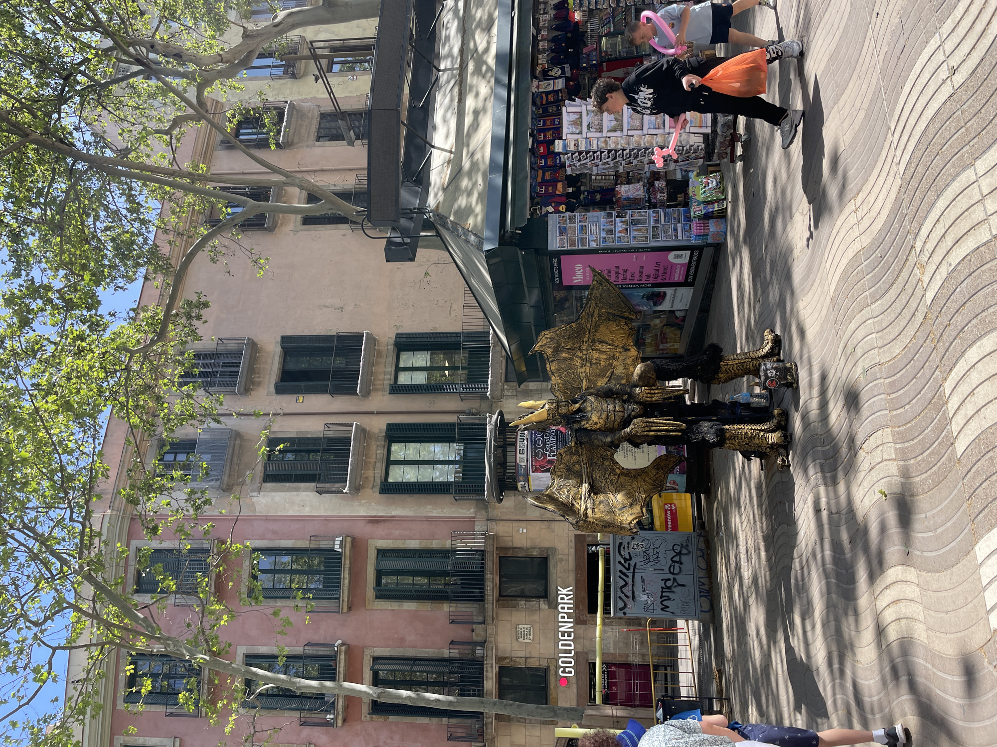 The image captures a street scene in Barcelona, Spain. The foreground features a person in a golden dragon costume, likely a street performer, positioned near a sign promoting Goldenpark. Behind the dragon, there's a newsstand/souvenir shop displaying various items. Passersby are visible, including individuals carrying balloons and shopping bags. The backdrop consists of a multi-story building with numerous windows, adding to the urban context. The street is paved with a distinct wavy pattern.