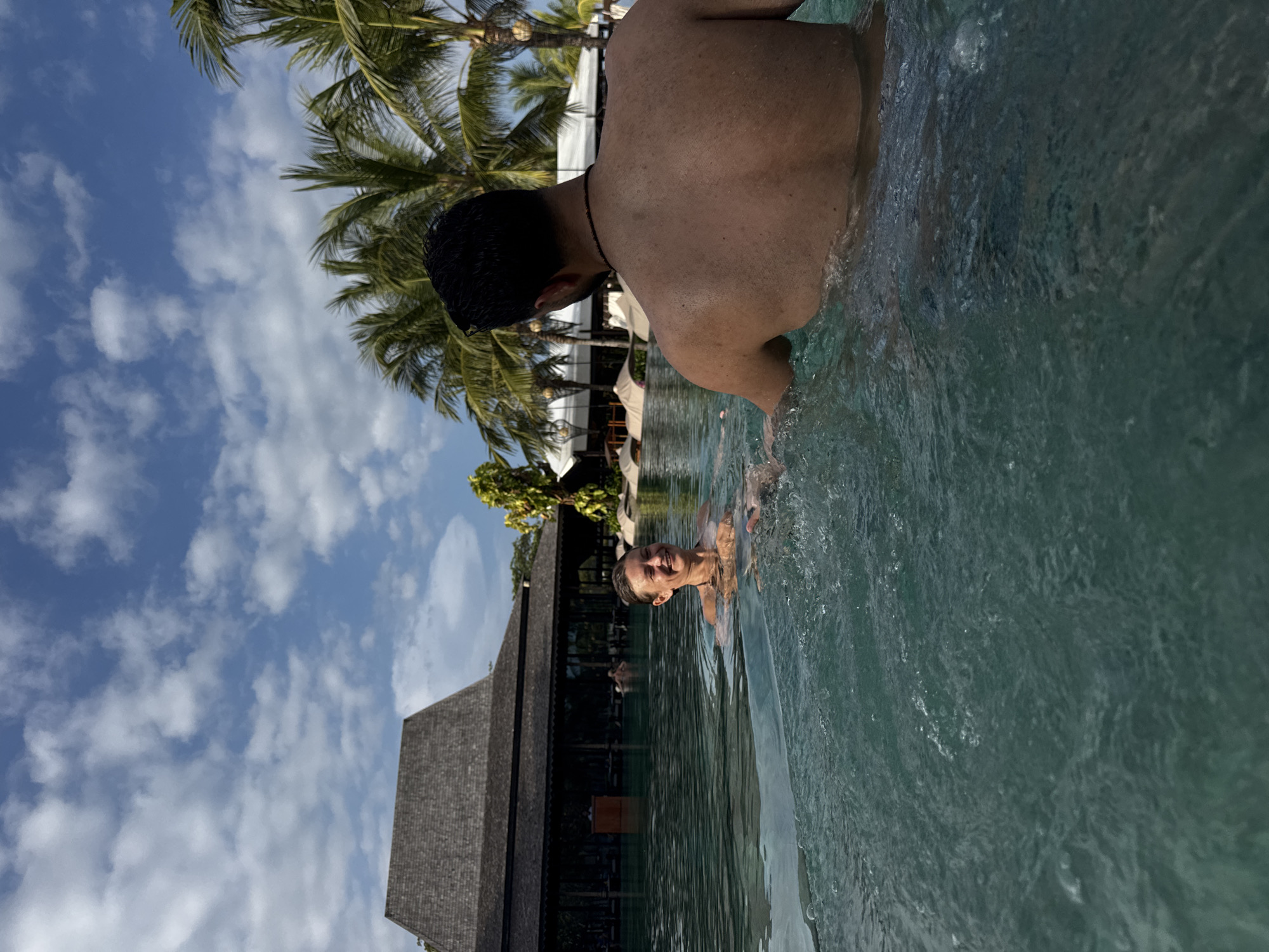 A person with dark hair and a bare back stands in a pool of greenish-blue water. Another person, a blonde with a fair complexion, is also in the water, looking towards the camera with a smile. The water is disturbed with ripples. In the background, a thatched-roof structure is visible, as are palm trees, a blue sky, and white clouds. The location is in Kuta Selatan, Indonesia.