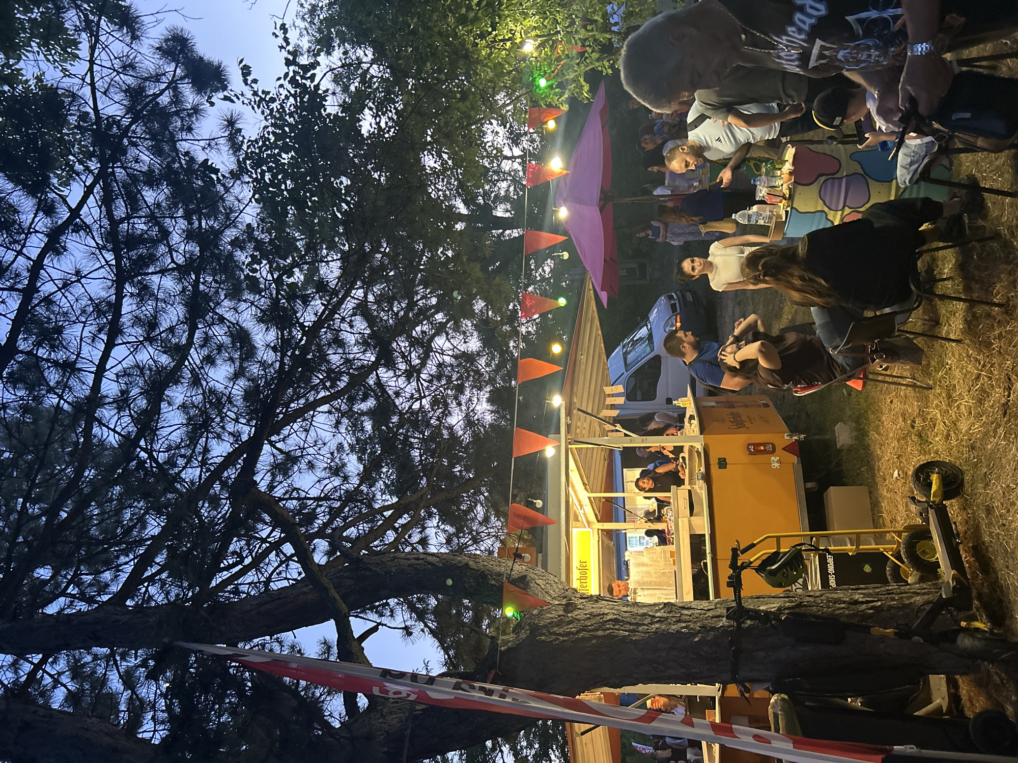 A group of people are gathered outdoors in the evening, possibly at an event or market. A food stand with a yellow awning displays the text "Schärferhofer" and is illuminated by string lights and orange triangular flags. Several individuals are visible behind the counter, appearing to serve customers. In the foreground, a few people are seated at tables, engaged in conversation and consuming drinks. To the left, a tree stands prominently with a red and white striped flag attached to it. A yellow scooter is parked near the tree. In the background, more trees and foliage create a natural setting. The overall atmosphere suggests a casual and lively social gathering.