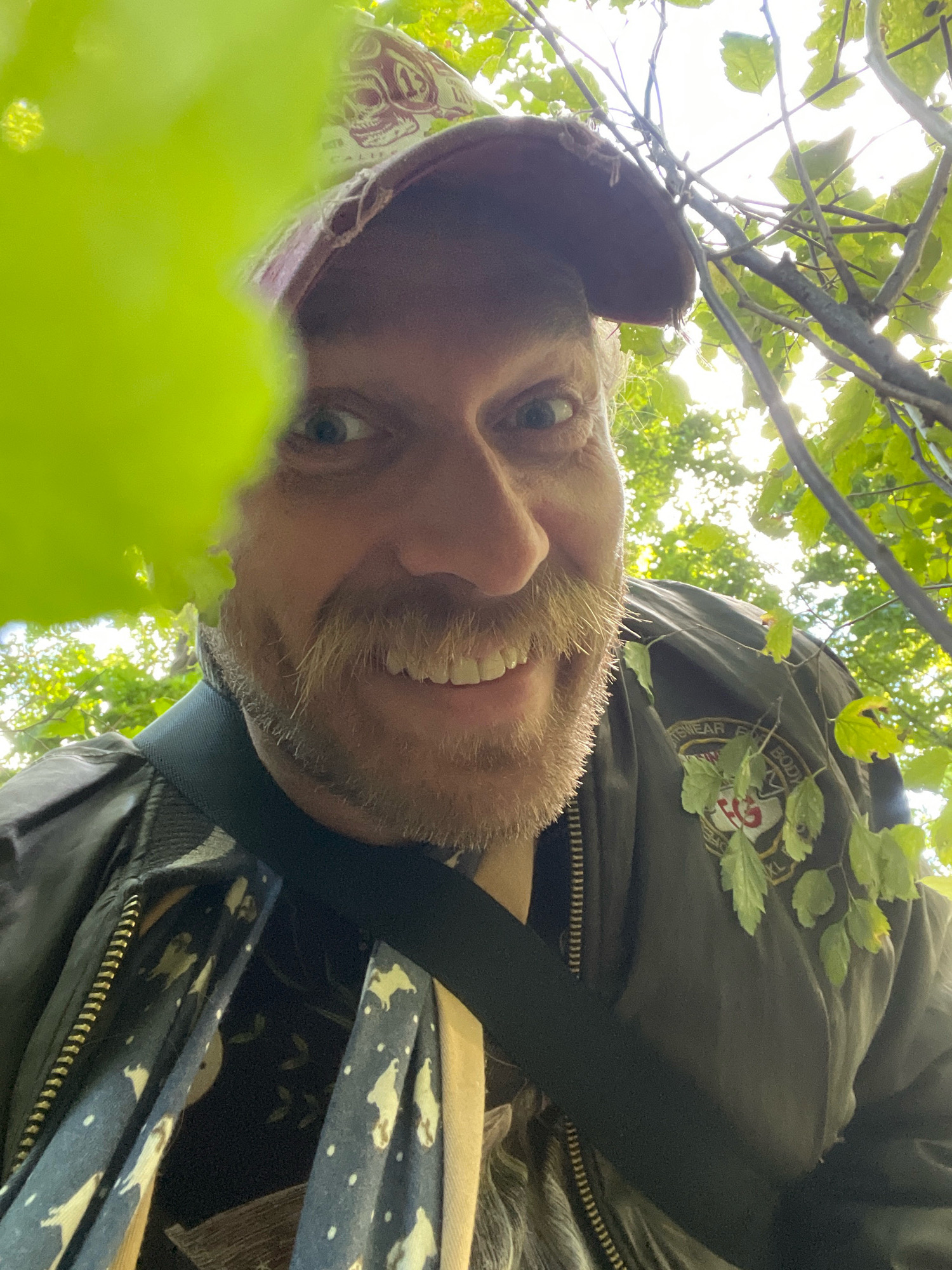A low-angle, close-up selfie shows a Caucasian man with light skin, a prominent mustache, and a wide smile, looking directly at the camera. He is wearing a dark green bomber jacket with patches on the left shoulder, a dark patterned scarf around his neck, and a camouflage baseball cap adorned with a skull design. The man is partially obscured by bright green leaves in the foreground on the left side of the frame. The background consists of dappled sunlight filtering through green foliage and tree branches, suggesting an outdoor setting. The image does not provide any visual cues that would allow for the identification of a specific location within Saint Petersburg, Russia.