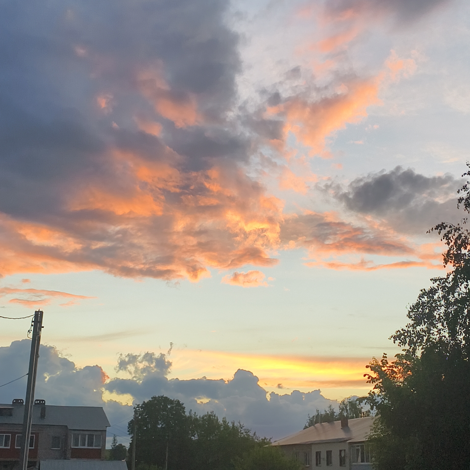 The image shows a sky at sunset in Фоки, Russia. The sky is filled with clouds, some of which are dark grey and others are illuminated with orange and pink hues from the setting sun. In the lower portion of the image, there are residential buildings with trees in front of them. A utility pole is visible on the left side of the frame. The scene is peaceful and indicative of a quiet evening in a residential area.