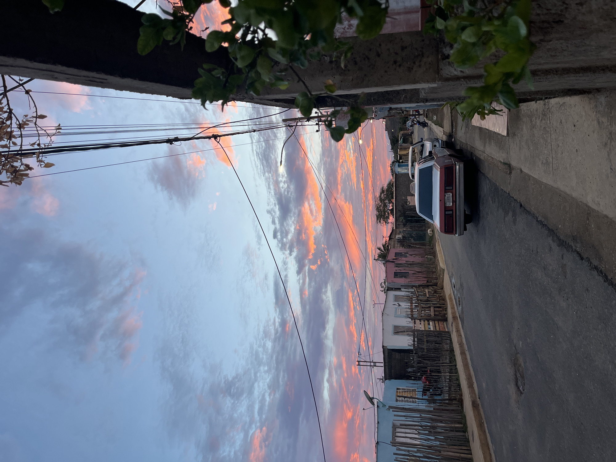 In Juan Griego, Venezuela, a street scene unfolds under a dramatic sky painted with hues of pink and orange as the sun sets. The foreground features an asphalt road with visible potholes, bordered by a concrete sidewalk. A pale gray wall, partially covered by lush green foliage, occupies the right side of the frame. Parked along the sidewalk are several vehicles, including a red and white sedan with a license plate reading "02NA8LO," and other cars further down the street.
On the left side of the road, a row of modest, brightly colored houses stands. The houses are constructed from various materials and are fronted by wooden fences. A red motorcycle is visible behind one of the fences. Overhead, a complex network of electrical wires crisscrosses the sky, supported by poles. The sky itself is a dominant feature, filled with wispy clouds tinged with the vibrant colors of sunset, transitioning to a pale blue higher up. The overall impression is one of a residential area at dusk, bathed in the warm glow of the setting sun.