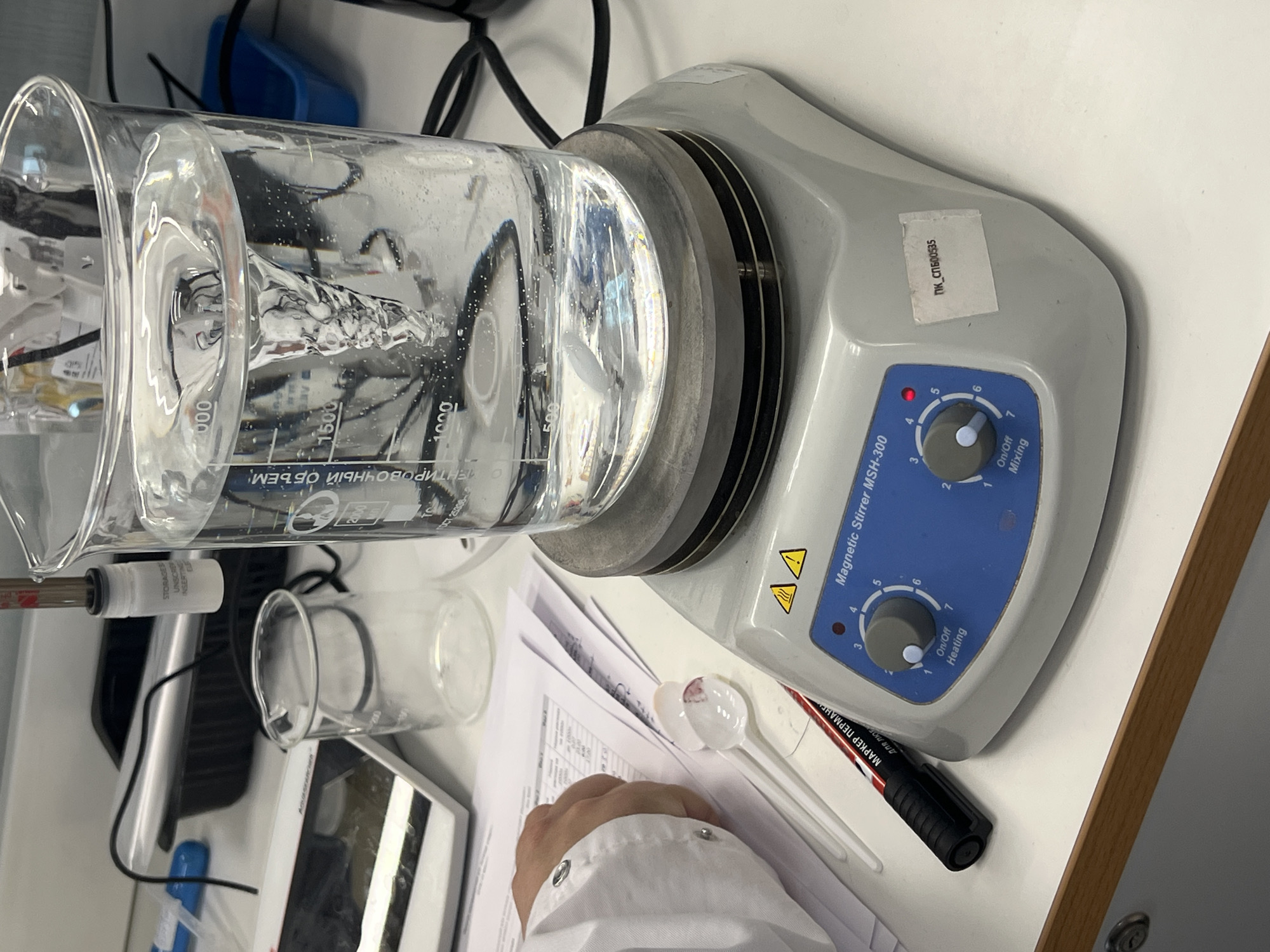 The image presents a laboratory setting, with a focus on scientific equipment and materials. A large graduated beaker, filled with a clear liquid, is positioned on a magnetic stirrer MSH-300. The magnetic stirrer has two knobs, one for mixing and the other for heating. The mixing knob is set to the "on/off" position, while the heating knob is set between 4 and 5. There is a white label on the stirrer with the text "ПК-СПБ00535".
Next to the beaker is a smaller graduated cylinder and a sheet of paper covered with text. There is a white plastic spoon with some red residue on it. A black marker is near the stirrer. A hand in a white lab coat is visible near the bottom of the image.
Based on the Cyrillic text visible on the equipment and materials (e.g., "МАРКЕР ПЕРМАНЕ", "МЕНТИРОВОЧНЫЙ ОБЪЕМ"), and the label on the magnetic stirrer, it can be inferred that the laboratory is located in a Russian-speaking region. However, there is not enough information in the image to determine the exact location in the city of Novosaratovka, Russia.