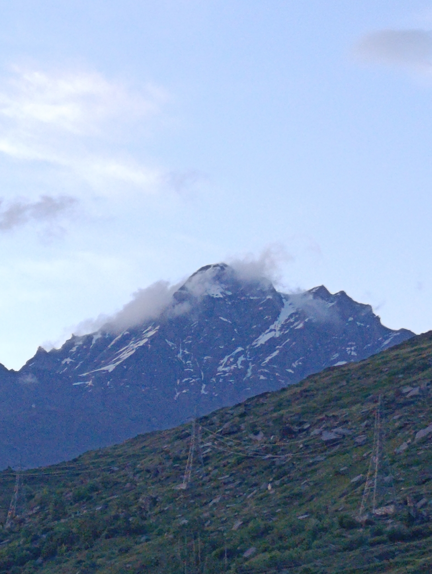 The image captures a majestic mountain peak partially shrouded in clouds, set against a pale blue sky with scattered wisps of white clouds. The mountain's slopes are rugged, with patches of snow visible, particularly on the upper reaches. In the foreground, a steep, rocky hillside is covered with dense green vegetation and scattered stones. Several tall, lattice-like electricity pylons are visible on this hillside, supporting power lines that stretch across the landscape. The overall impression is one of a remote, high-altitude natural environment, possibly in a mountainous region of India.