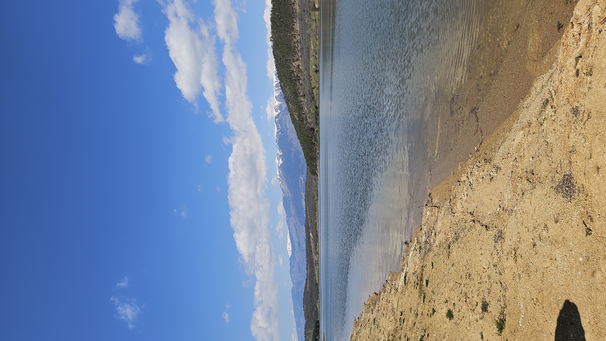 The image depicts a landscape with a lake, mountains, sky, and shoreline. On the left side of the image, the sky is blue with scattered white clouds. In the middle, a mountain range is visible, with some snow on the peaks. A line of trees runs along the base of the mountains, near the lake. The lake is present in the middle of the image, with a visible shoreline on the right. The shoreline is sandy and slopes downward toward the water.
Given the location of Domlyan, Bulgaria, and the terrain depicted, this could be a lake and mountain landscape in the region.
