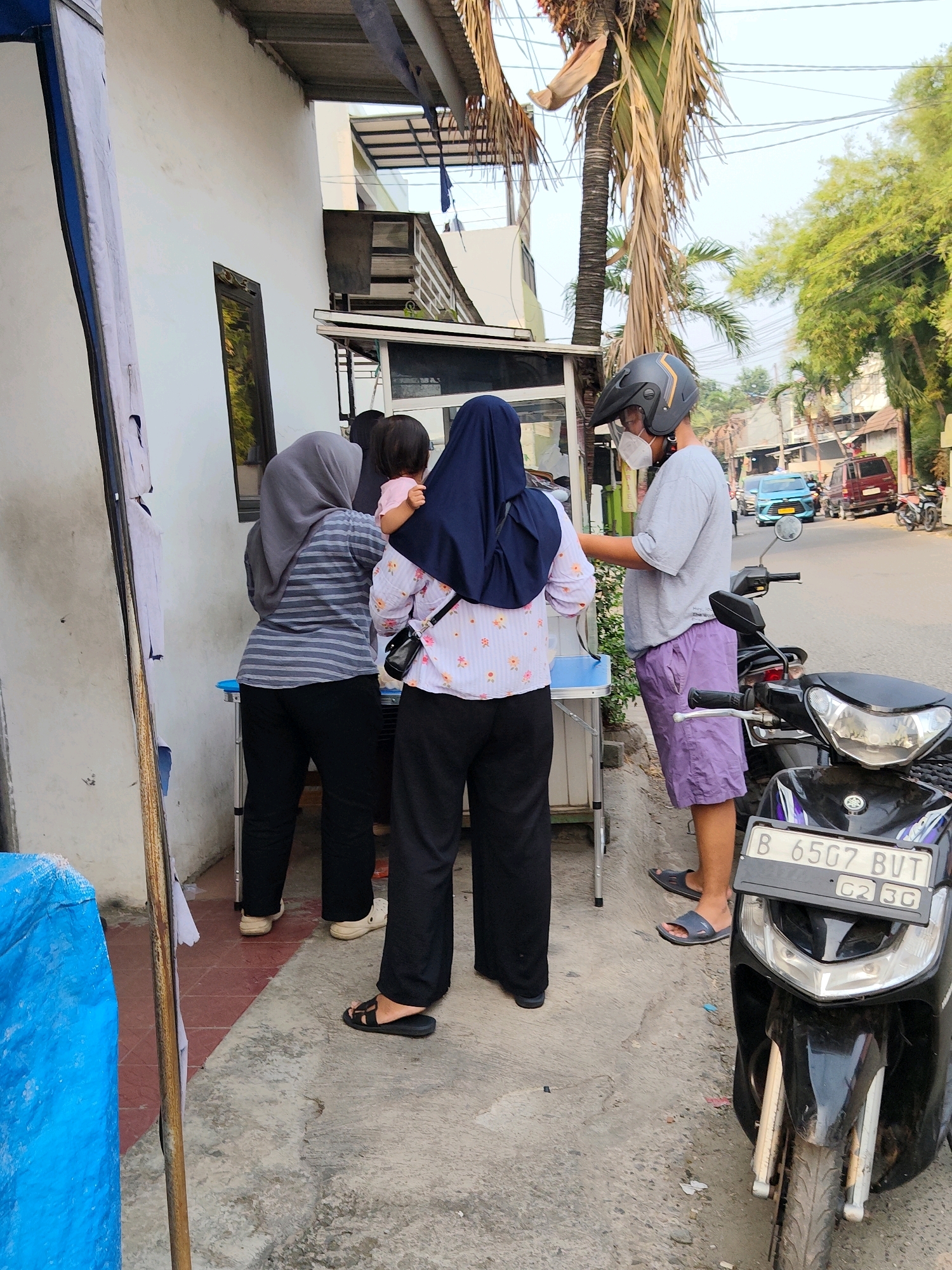 In Bekasi, Indonesia, on a street with a building on the left and a road on the right, a group of people are gathered near a table. A man wearing a helmet and a face mask is standing, facing the table. To his left, a woman wearing a dark blue hijab and a floral patterned shirt is holding a baby on her shoulder. Another woman in a grey hijab and a striped shirt is also present, facing the table. A black motorcycle is parked next to the man. The environment appears to be an outdoor setting, possibly a market or a roadside stall, with a palm tree visible in the background.