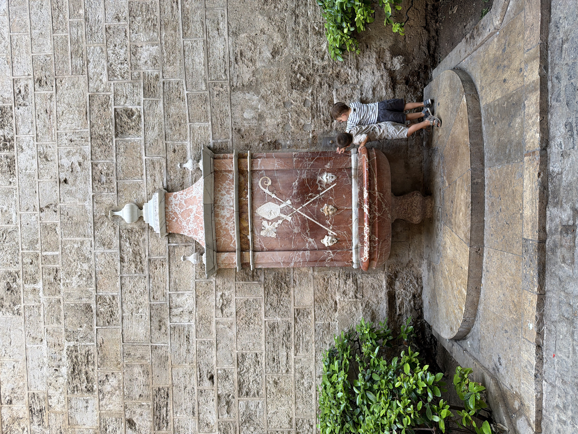 Two young boys are standing at a red marble fountain against a stone wall. One boy, wearing a striped shirt and shorts, is reaching out to the fountain. The other boy, wearing a grey shirt and shorts, is leaning against the fountain. The fountain has a carved design of crossed staffs and three faces. Green bushes are on either side of the fountain. The ground in front of the fountain is paved with stone tiles.