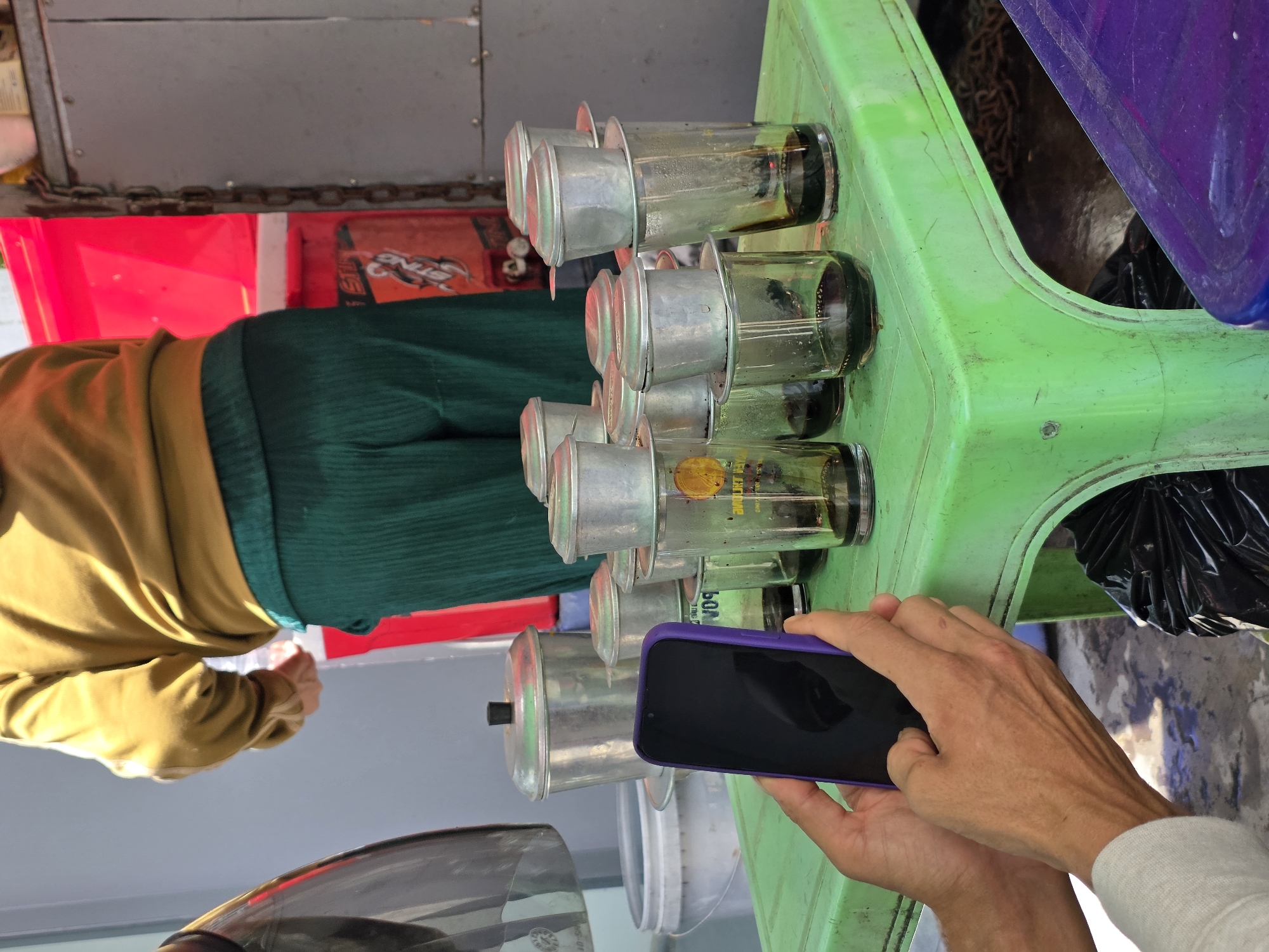 In Binh Thuan Ward, Vietnam, a person is holding a purple smartphone in their hands, partially obscuring a row of Vietnamese coffee filter setups. These setups consist of small metal filter devices stacked on top of clear glass cups, each containing a dark liquid, presumably coffee. The cups are arranged on a bright green plastic table. Behind the table, a person wearing a mustard-yellow sweatshirt and dark green pleated pants is visible, seemingly engaged in preparing or serving something. To the right, a large red container and a metal chain are noticeable. The ground is concrete, with a black trash bag visible beneath the table.