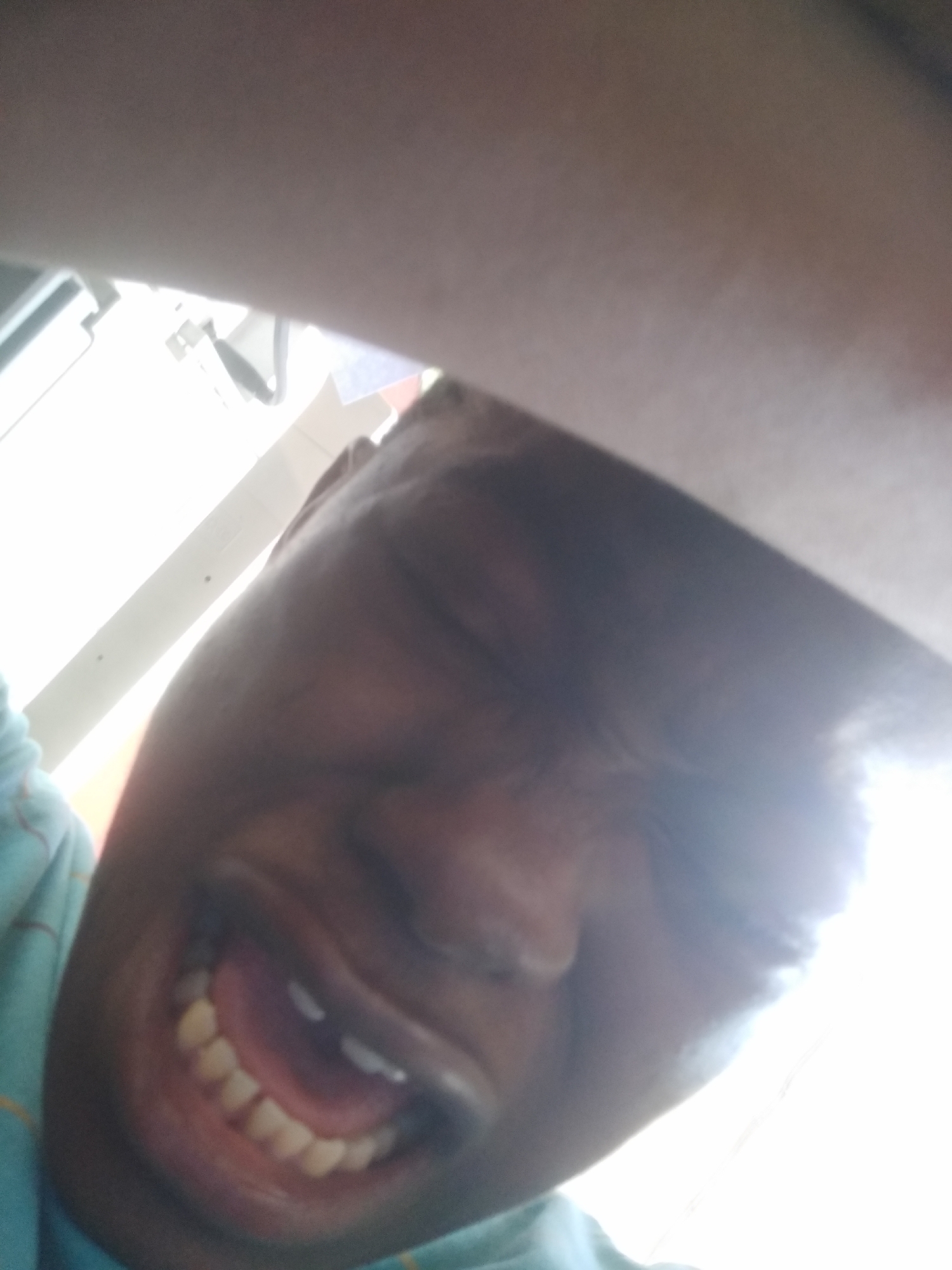 A close-up, low-angle shot captures a person's face contorted in an expression of apparent discomfort or distress. The person's eyes are tightly shut, and their mouth is open, revealing their teeth. The surrounding environment appears to be indoors, with a bright light source illuminating the scene. The location is Shangisha, Nigeria, but there are no details in the image that confirm this or further describe the location.