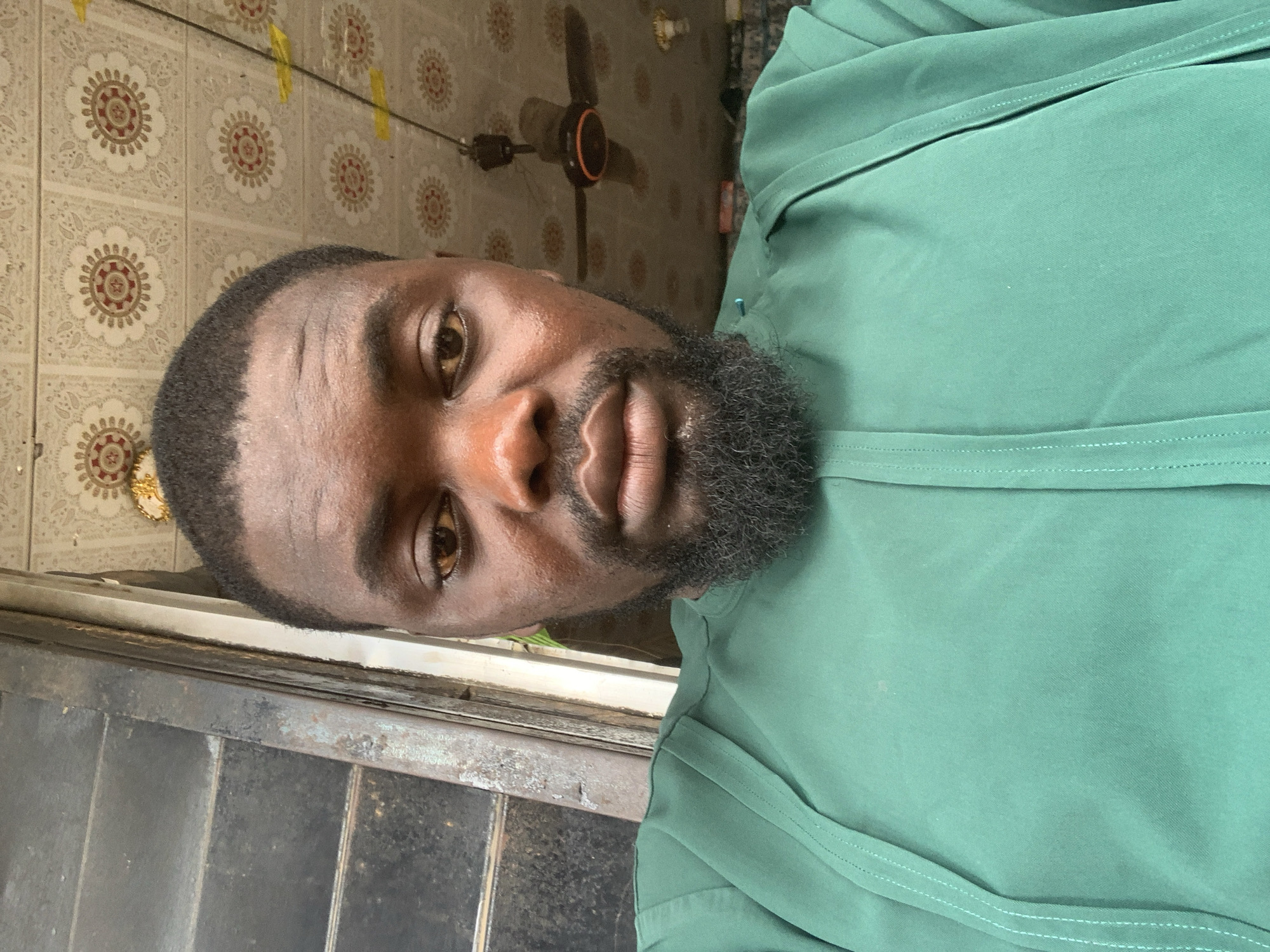 A close-up, head-and-shoulders selfie is taken by a Black man wearing a green traditional garment. The man has dark skin, a short haircut, and a full beard. His eyes are looking directly at the camera. The background of the image shows a ceiling with floral-patterned tiles and a ceiling fan. To the left of the man, a window with dark, slatted blinds is visible. The overall lighting suggests it is daytime, possibly indoors or under a covered area. The man appears to be in a residential or communal space. There is no specific identifiable information within the image that pinpoints the location to Shagamu, Nigeria.