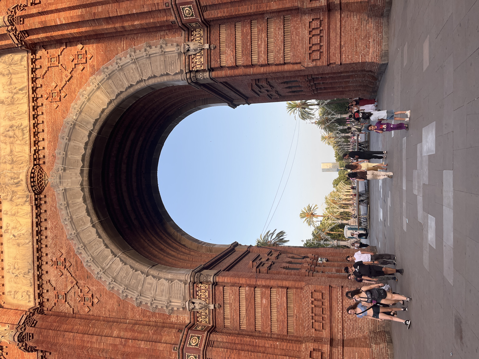 The image shows the Arc de Triomf in Barcelona, Spain. The monumental arch is constructed of red brick and features ornate carvings and a frieze depicting allegorical figures. The archway is open, revealing a clear blue sky and a pathway lined with palm trees. Several people are walking in the foreground and midground, some appearing to be tourists. The scene is bathed in warm, late afternoon sunlight.