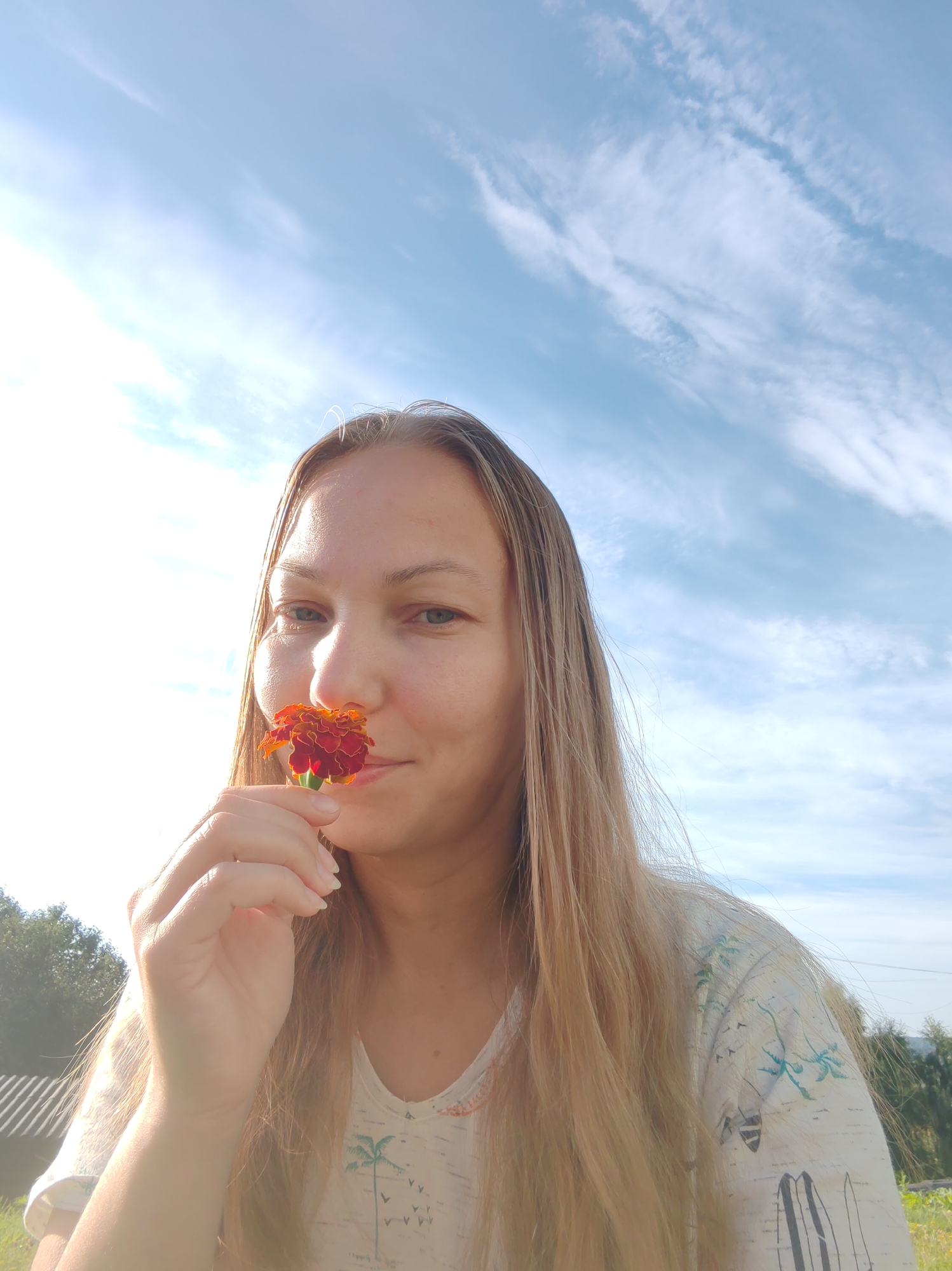 A young Caucasian woman with long blonde hair and fair skin is captured in a close-up, outdoor, daytime selfie. She is holding a small, red and orange marigold flower up to her nose, as if smelling it. Her eyes are open, and she has a slight smile on her face. She is wearing a light-colored t-shirt with a palm tree and bird print. The background consists of a bright blue sky with wispy white clouds, suggesting it is a sunny day. To the left of the frame, a green tree line and a striped structure, possibly a fence or roof, are visible. The overall impression is a serene and pleasant moment outdoors. The location is specified as Chukhlem, Russia.