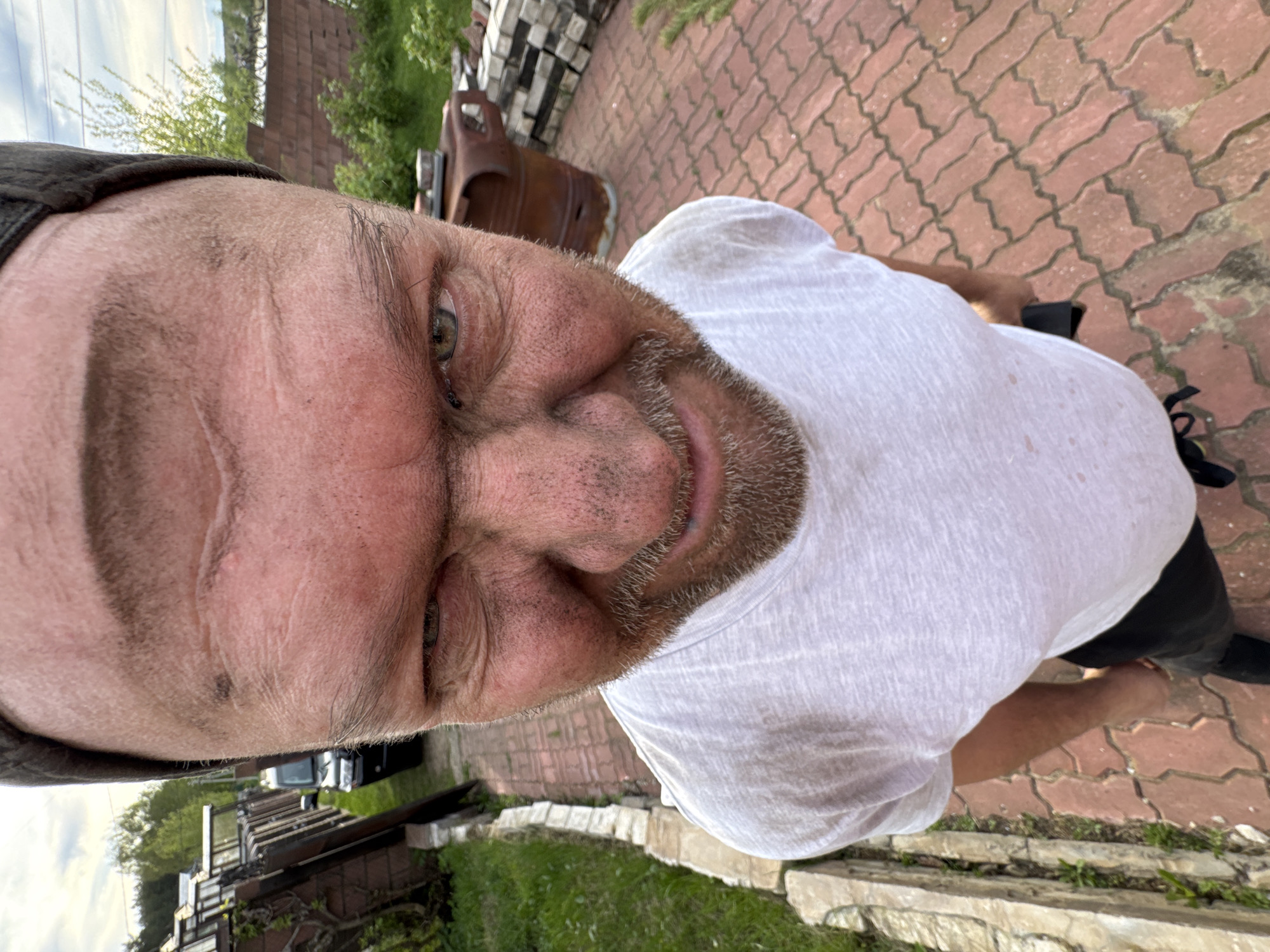 A selfie shot of a man with a white shirt and beard is standing on a red brick surface. The photograph is taken outdoors, in what appears to be Bol'shoye Kozino, Russia. In the background, there are stacks of stones and what may be an old vehicle part. A partial view of a house and green foliage suggests a residential or semi-rural setting.