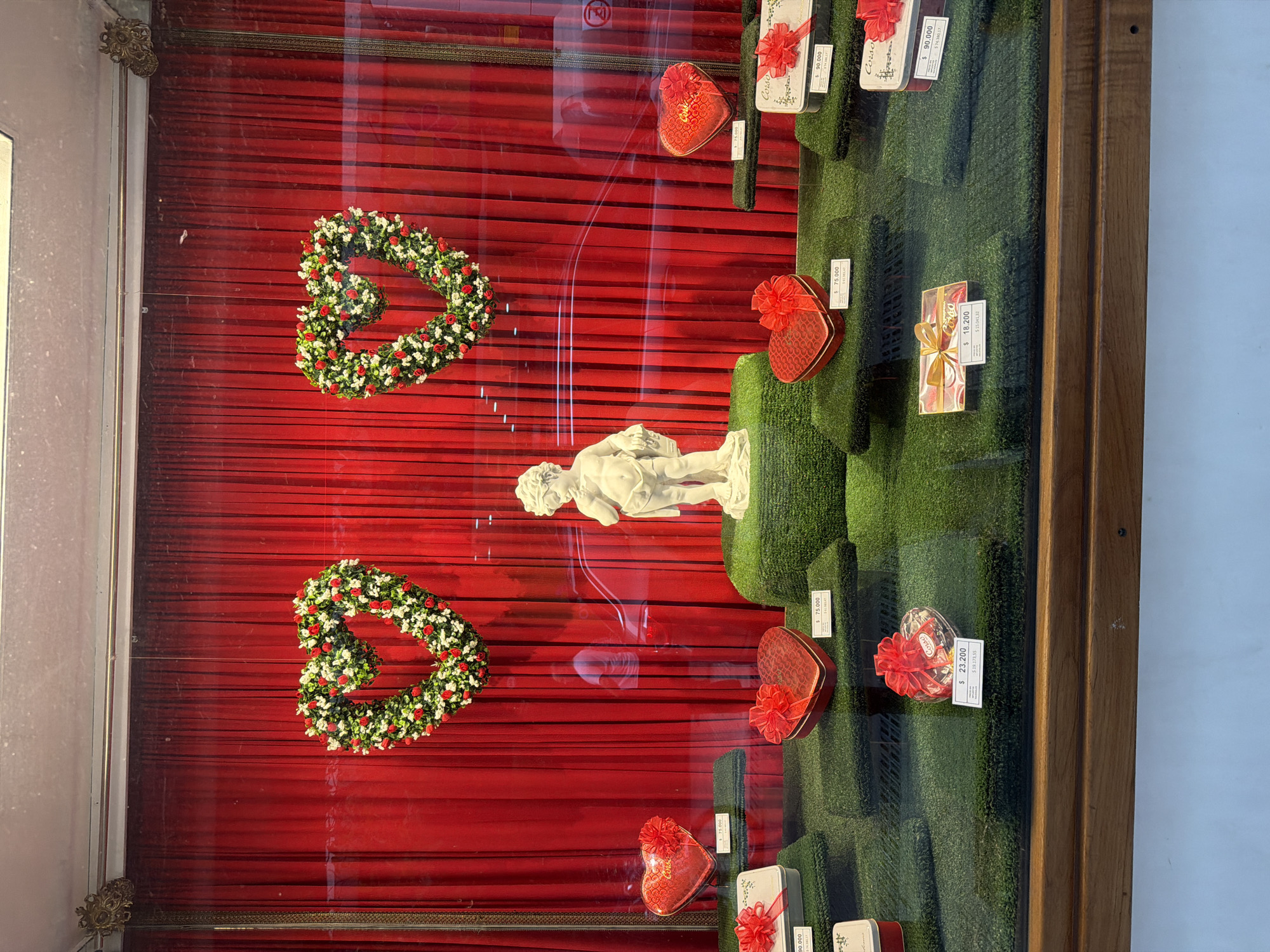 The image displays a shop window, likely in Buenos Aires, Argentina, filled with items set against a vibrant red draped background. The window appears to be decorated for a romantic occasion, such as Valentine's Day.
**Subjects:**
The primary subjects are various gift items presented on tiered, grass-like green platforms. These include several heart-shaped gift boxes, predominantly red with red bows, as well as some rectangular or square gift boxes, also adorned with red bows. A prominent white statue resembling a cherub or cupid stands in the lower-center of the display. Two large, heart-shaped wreaths, made of red, white, and green artificial flowers, hang against the red background.
**Activity or event taking place:**
The scene depicts a retail display, showcasing products for sale. The overall theme, indicated by the hearts, cupid, and gift boxes, suggests a focus on gifts for a romantic holiday or event.
**Notable details:**
The display is contained within a wooden-framed window, and reflections on the glass obscure some details but indicate an outdoor environment beyond. The bright lighting suggests it's daytime. The presence of currency on the price tags, likely Argentine Pesos given the location context, highlights the commercial nature of the display. The image is oriented sideways, with the display appearing to be a vertical window pane.
**Visible text:**
Several price tags are visible, showing amounts such as "$ 90.000" (appearing multiple times), "$ 23.200", "$ 19.173,55", "$ 75.000", "$ 18.200", "$ 15.041,32", and "$ 74.380,17". The brand name "Corso" also appears on some tags or packaging.