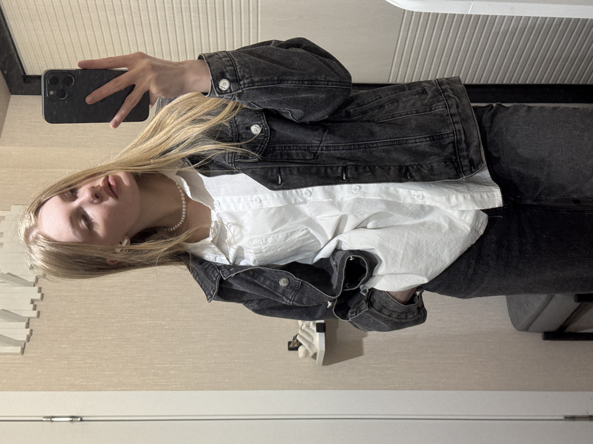 A young woman with long blonde hair is depicted in a mirror selfie, holding a black smartphone with a triple camera module in her right hand. The image is rotated 90 degrees counter-clockwise, showing her upright as she captures the moment. She is dressed in a dark grey/black washed denim jacket worn over a loose, white button-down shirt, and matching dark grey/black jeans. A delicate white pearl necklace is visible around her neck, and a white earbud is nestled in her right ear. Her expression is neutral, with her gaze directed towards the camera. The background reveals an indoor setting with a light, neutral-colored wall featuring a subtle horizontal textured pattern. Above her, white horizontal blinds or a similar paneling are visible. To her left, near her waist, a light-colored object rests on a surface, and in the lower right, a grey cushioned chair with a black frame is partially visible. No discernible text appears in the image. The scene is set indoors, within a room in Perm, Russia.