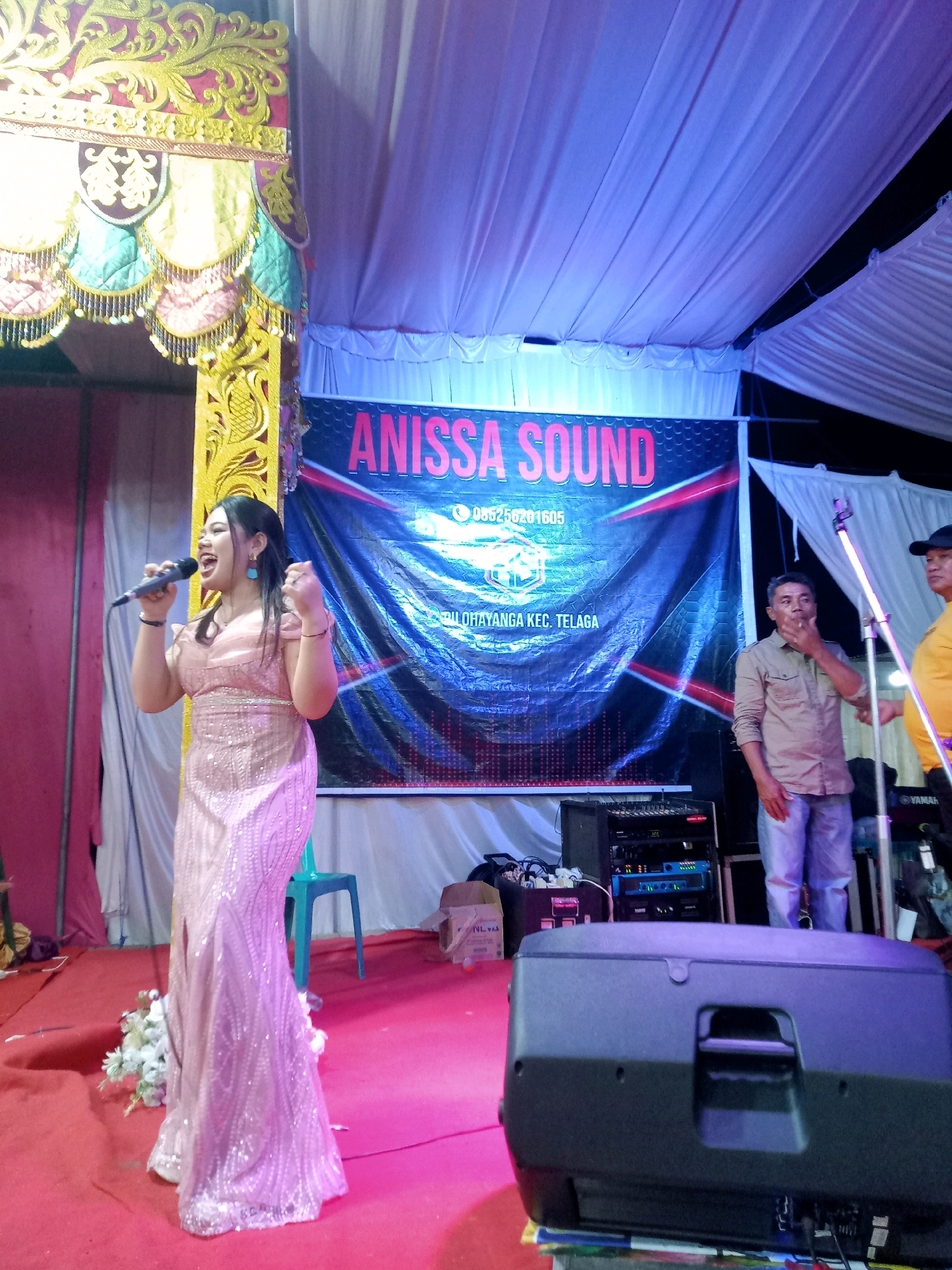 A full-shot, eye-level, indoor candid photograph captures a young woman in a sparkly pink floor-length dress performing on a stage in Mongolato, Indonesia. The woman is positioned to the left of the frame, holding a microphone in her right hand and gesturing with her left. She is looking upwards and to her left, with her mouth open as if singing. The stage is covered in a red carpet, and behind her is a large banner with pink lettering that reads "ANISSA SOUND" above a phone number and location information. The background consists of a draped ceiling illuminated with blue lights and various pieces of audio equipment, including speakers and a mixer board. To the right of the woman, two men are standing, one of whom is holding his hand to his chin and looking towards the audience. The overall environment suggests an outdoor event or performance space covered by a tent.