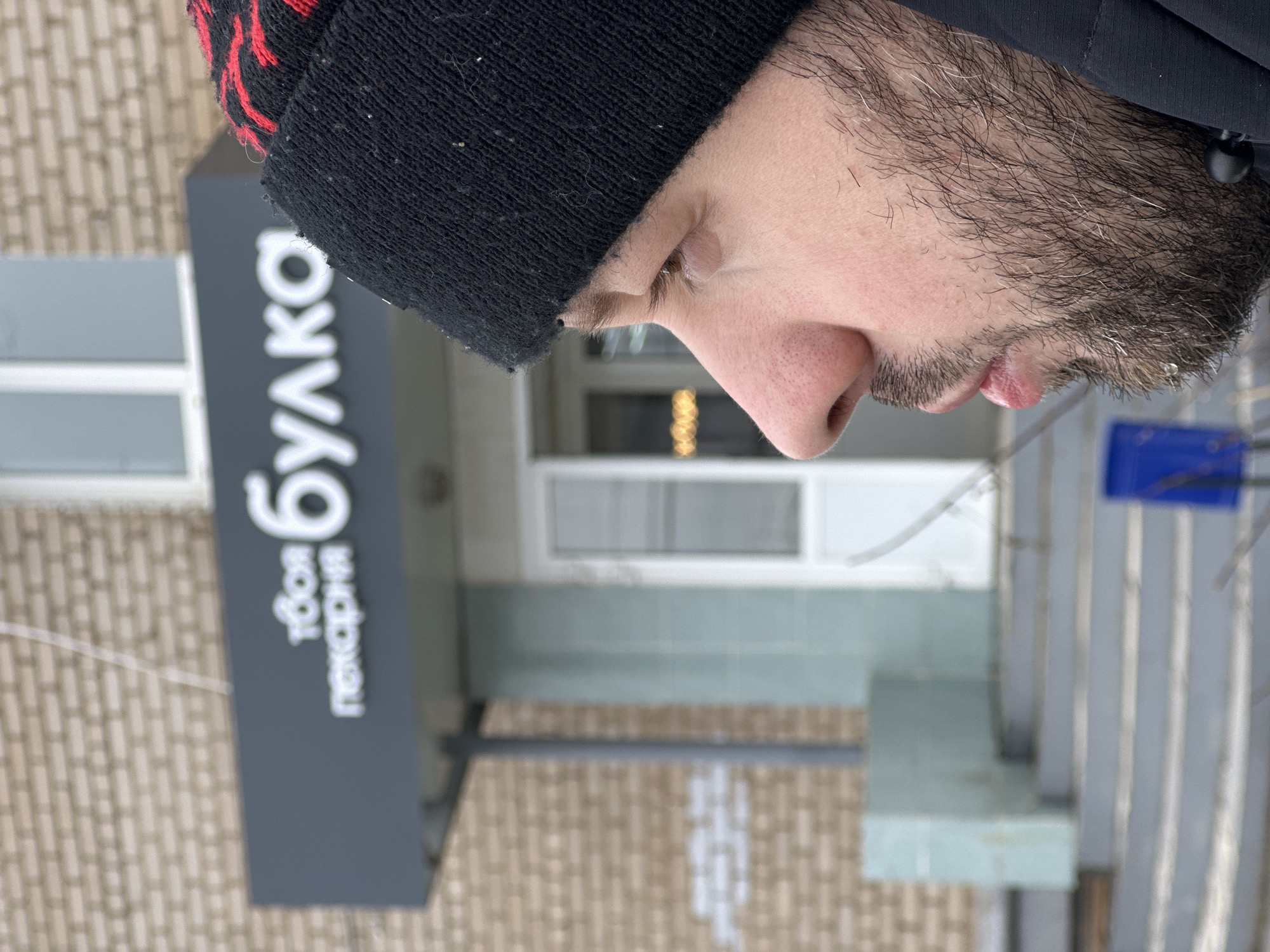 A man with a black and red beanie stands in front of "Твоя пекарня Булка" (Your Bulka Bakery) in Chaikovsky, Russia. He is in the foreground, slightly out of focus, while the bakery storefront is in the background. The bakery sign is gray with white lettering. The building behind the sign is beige brick. The scene has a muted, slightly cold tone, possibly indicating an overcast day.