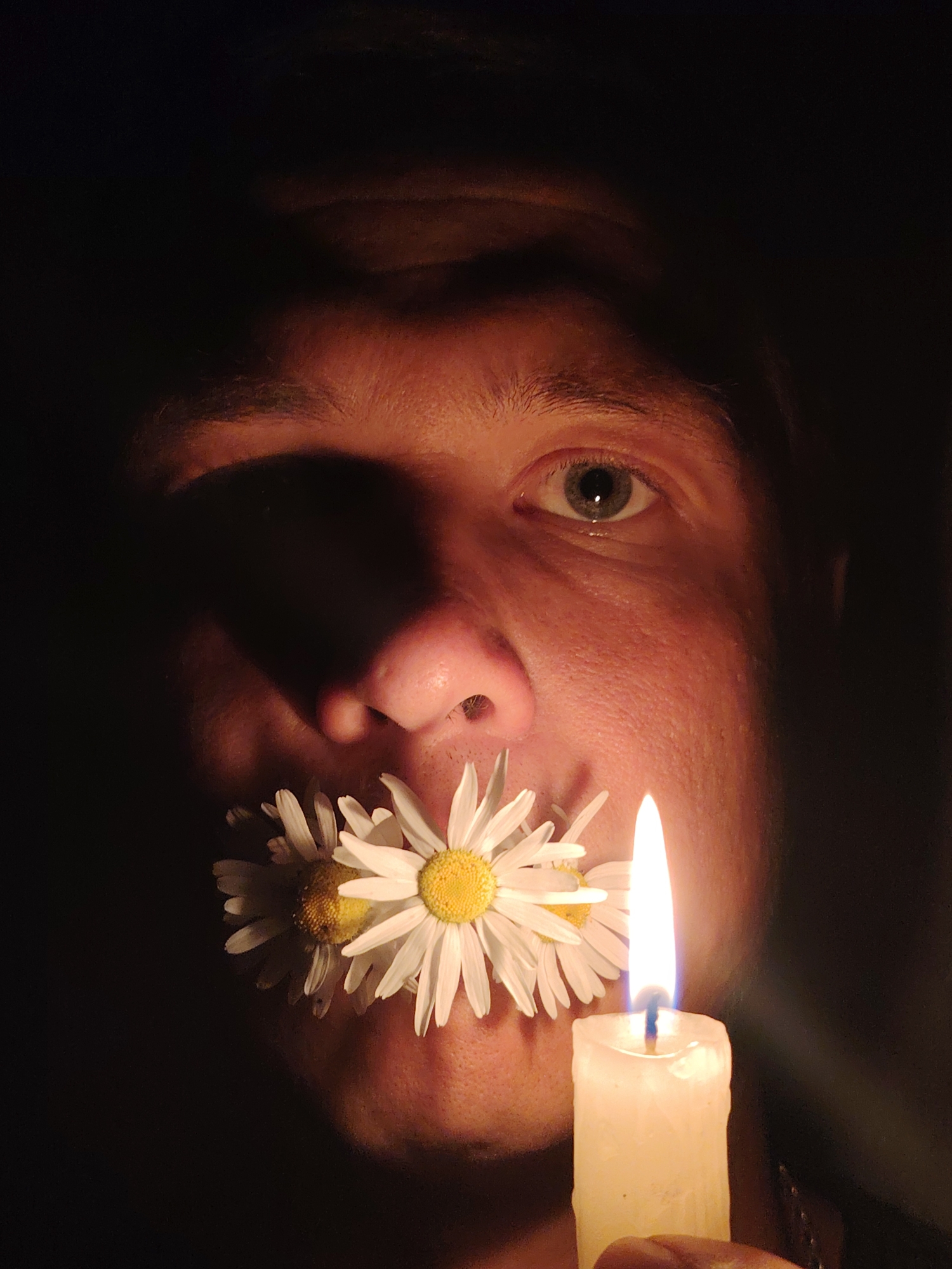 The image is a close-up portrait of a person's face, illuminated by candlelight. The person's mouth is obscured by two white daisies. Their left eye is clearly visible, with a grey iris. The right side of their face is in shadow. A lit white candle is positioned to the right of their face, casting a warm glow. The background is dark and indistinct. There is no information in the image to suggest a specific location in the city of Chaikovsky, Russia.