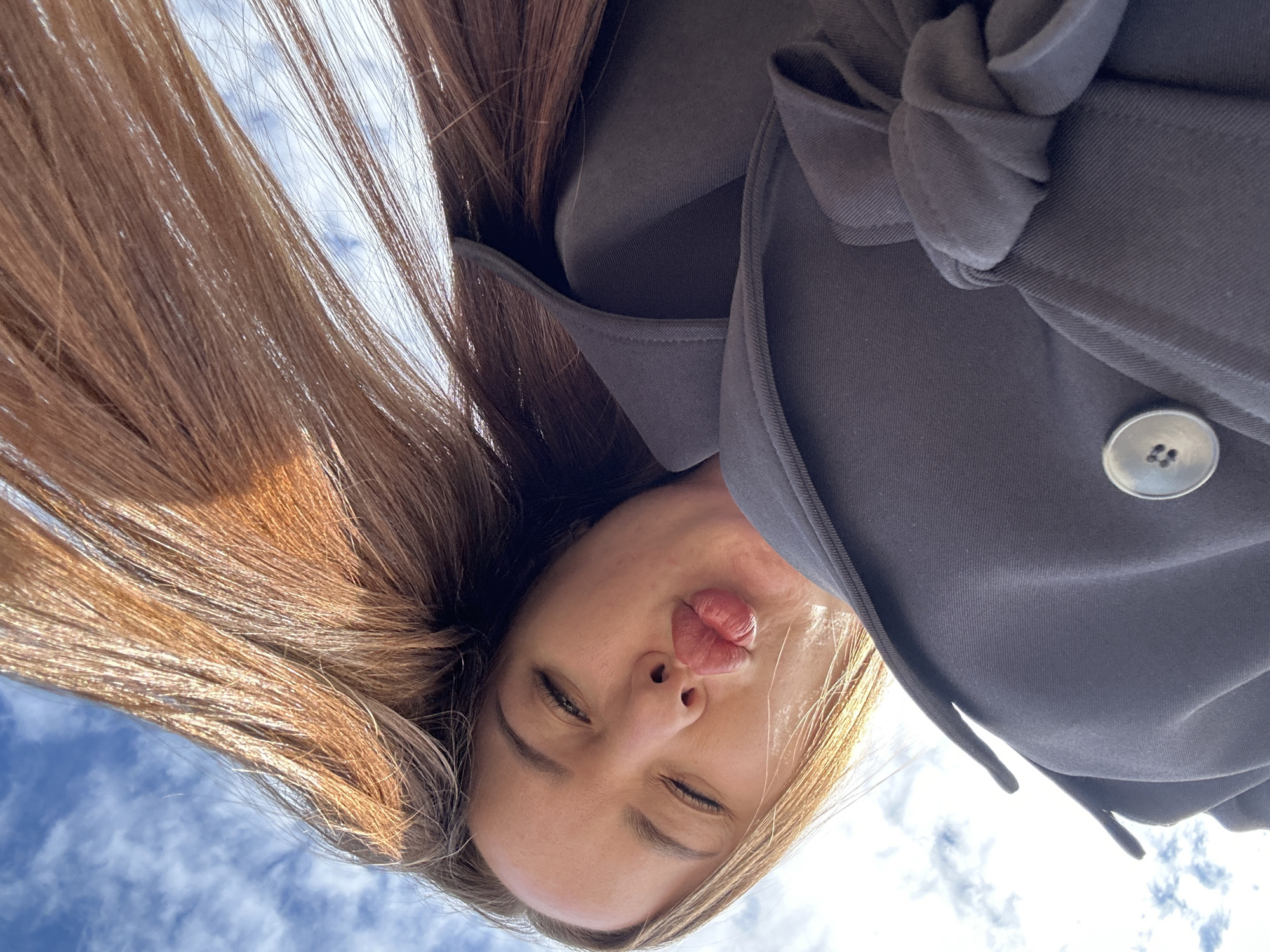 An outdoor, low-angle portrait captures a single individual in the city of Noviy, Russia, positioned above the camera's perspective. The subject, appearing female, exhibits long, light brown to auburn hair fanned out upwards and to the sides. Her face is angled downward, with lips pursed forward in a distinct "kissing" gesture. The left eye is closed, while the right eye remains partially open. She is dressed in a dark grey or charcoal-colored outer garment, possibly a coat or jacket, featuring a visible light-colored, four-hole button on the lower portion and a matching fabric tie or knot on the right side of the frame. The background consists of a blue sky with scattered white clouds, indicating a daytime setting. No other individuals or environmental structures are present in the frame.