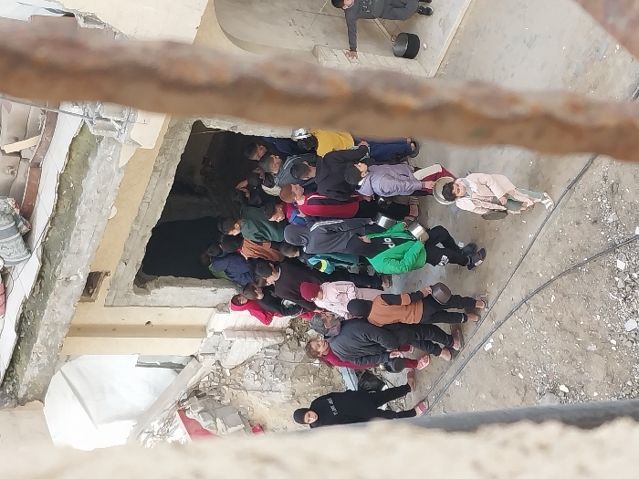 This image depicts a damaged urban environment in Khan Yunis, Palestinian Territory, viewed from an elevated perspective. A severely compromised, multi-story concrete building occupies the background, exhibiting extensive structural damage, exposed rebar, and large sections of broken concrete and rubble on its upper levels and immediate surroundings. The ground is composed of dirt and scattered debris, with dark linear objects, possibly cables, traversing the foreground.
Approximately 30 to 40 individuals, predominantly children and young adults, are congregated near a dark, irregularly shaped opening in the lower portion of the damaged building, which appears to be a breached wall or entrance. Many individuals in the crowd are holding metal pots, pans, and other containers, suggesting participation in a distribution or collection event, likely for food or water. People are observed moving into, out of, or queuing near this opening.
Specific subjects include: a woman in a black abaya and hijab standing to the far left; an adult male with a beard, wearing a dark jacket, walking leftward while carrying a pot; numerous children and adolescents, dressed in casual winter clothing such as hoodies and jackets, each holding a container; and a young child in a light-colored coat in the mid-foreground, carrying a flat metal object. On the far right, a young boy in a grey tracksuit stands with arms outstretched near a black pot on the ground. The scene conveys a collective gathering for resources within a setting of widespread structural damage.
