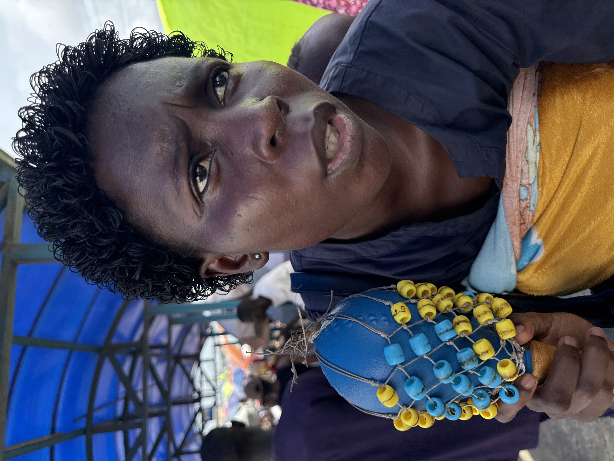 A dark-skinned person with curly hair, possibly in their late 20s to early 40s, is holding a blue musical instrument adorned with yellow and blue beads connected by a string net. They are wearing a dark blue top. The background shows blurry figures of other people and what appears to be a semi-enclosed structure with a blue roof. Based on the overall feel and known activities, the location is likely a public area or event in Lekki, Nigeria.