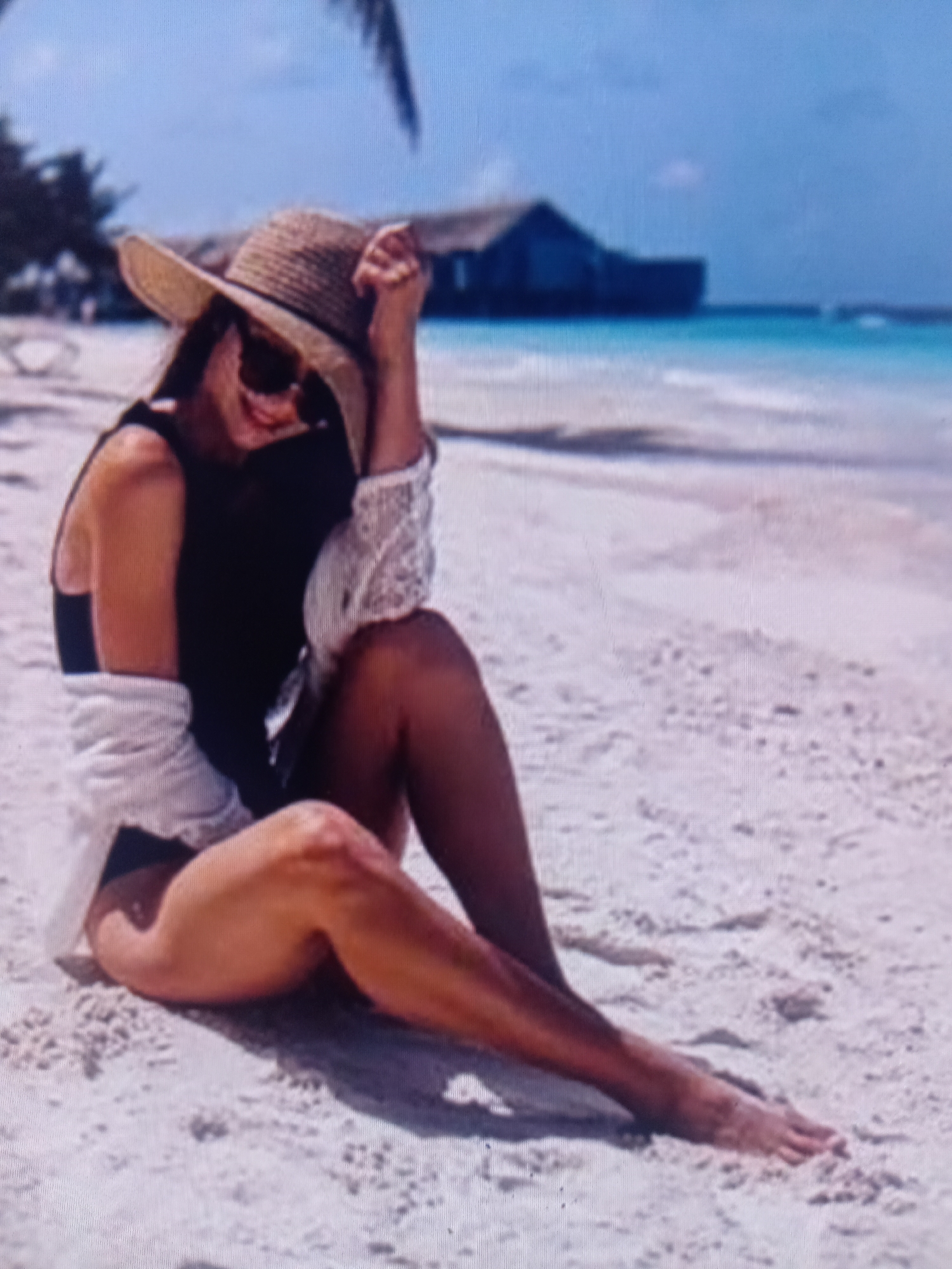 A person sits on a sandy beach, wearing a straw hat, sunglasses, and a black swimsuit. A white cover-up is draped over their shoulders. They are leaning back, with their legs extended, and their hand is raised to their hat. The person appears to be smiling or laughing. The background shows a turquoise ocean and a dark building on stilts. The sky is blue with some clouds. The overall scene suggests a tropical vacation or a day at the beach.