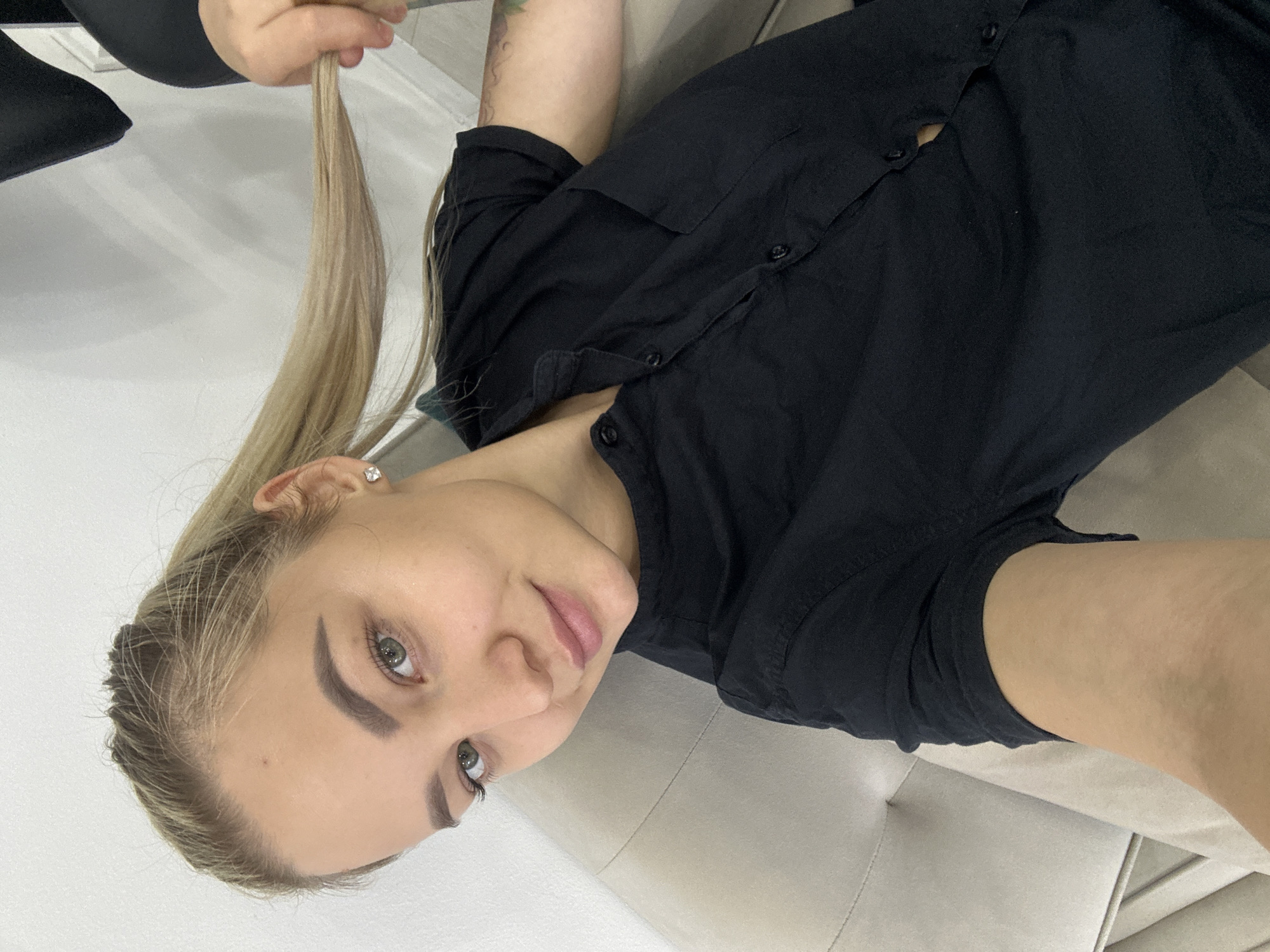 A woman with blonde hair, light skin, and green eyes is lying on a light-colored couch. She is wearing a black button-down shirt. Her right hand is holding a strand of her hair. The location is potentially Budva, Montenegro, but without more information, it is impossible to confirm the city.