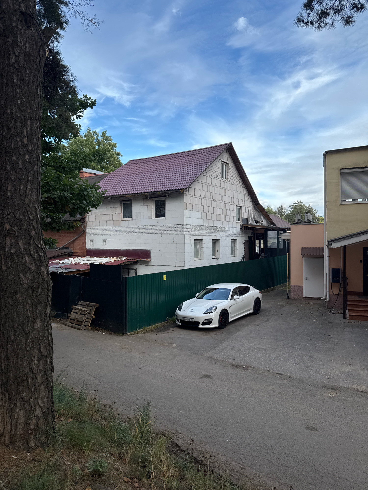 This is a street-level, medium shot of an urban scene in Novoivanovskoe, Russia. A white Porsche Panamera is parked on an asphalt road in front of a multi-story building under construction, made of white concrete blocks, with a burgundy metal roof. A green metal fence separates the road from the property. To the right of the construction site, a lighter-colored building with a grey roller shutter window is visible. A large tree trunk is in the foreground on the left side of the image. The sky is partly cloudy with a blue and white sky.