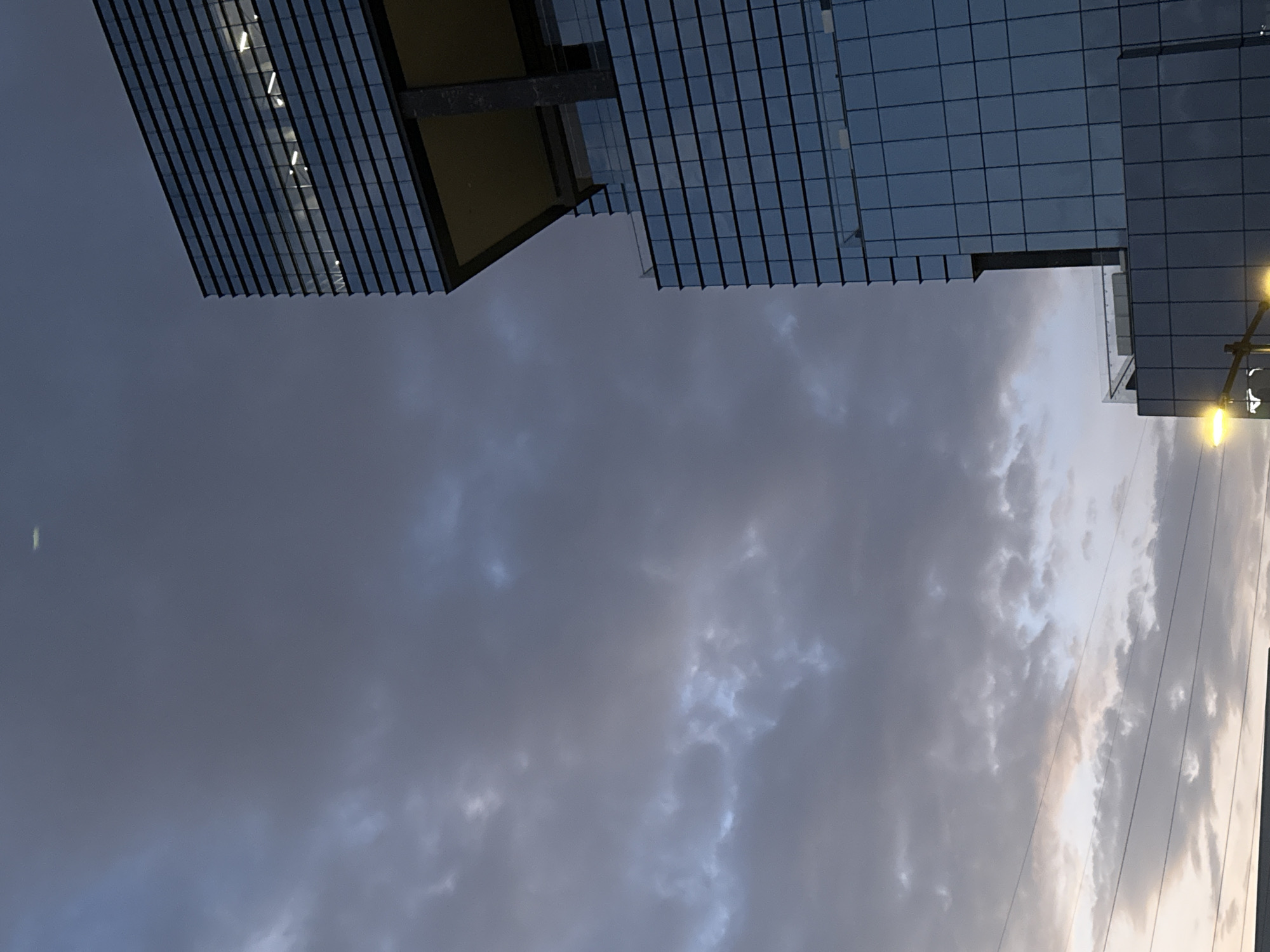 The image shows a low-angle view looking up at modern architecture under a cloudy sky, possibly taken in the city of Puebla, Mexico. Predominantly visible is the underside of a building with a grid-like facade, suggesting it's made of glass and steel. The sky is overcast, with the diffused light filtering through the cloud cover. Streetlights are partially visible on the right. Power lines stretch across the sky, adding to the urban setting.