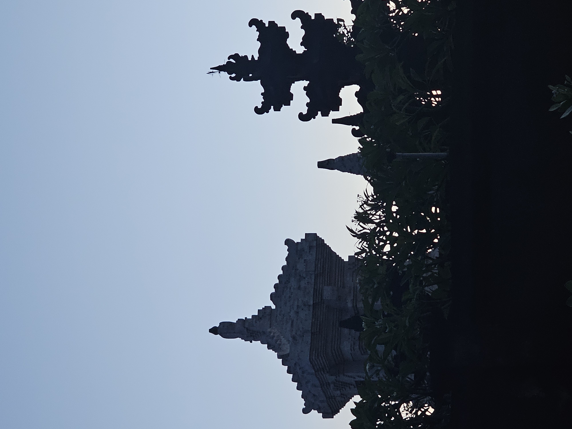 The image, likely taken in Denpasar, Indonesia, shows a silhouette of a multi-tiered stone structure, likely a Balinese temple or shrine, viewed from a low angle. The sky is a pale blue. The structure features intricate carvings and ornamentation, typical of Balinese architecture. Part of the structure is obscured by a dark mass, possibly a tree.